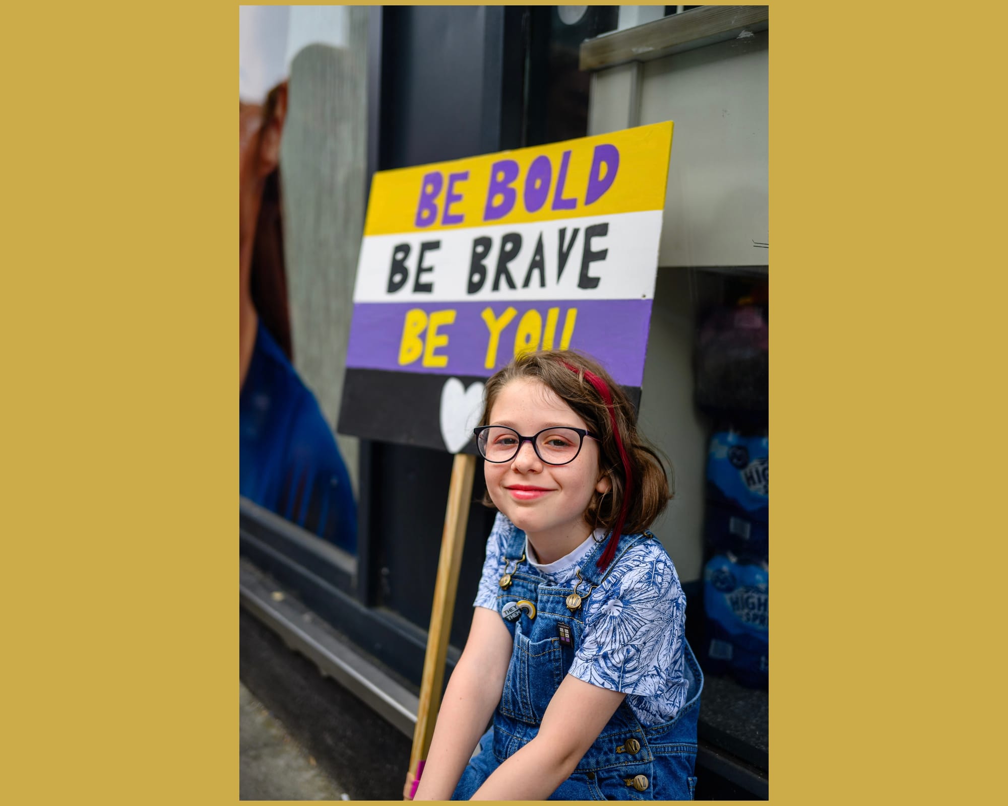 A young kid wears a "they/them" pronoun pin with red lipstick and a nice smile. Behind is a sign saying "Be bold. Be brave. Be you." in the non-binary flag colours.