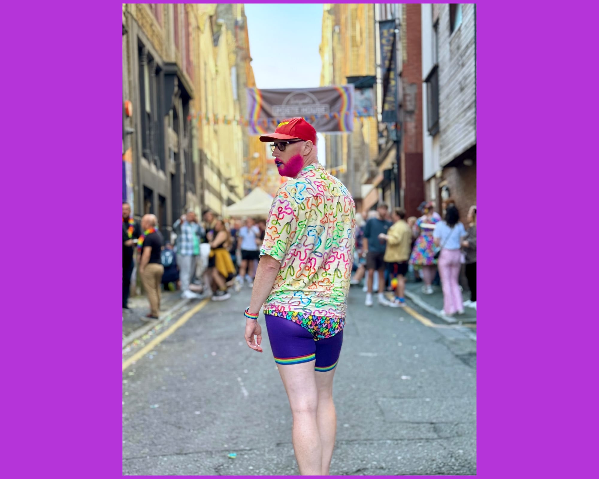 Non-binary person wearing a shirt with rainbow squiggles on. Below they are wearing purple bicycle tights with rainbows at the end. Over that is a pair of rainbow heart briefs. They also have a pink beard. 