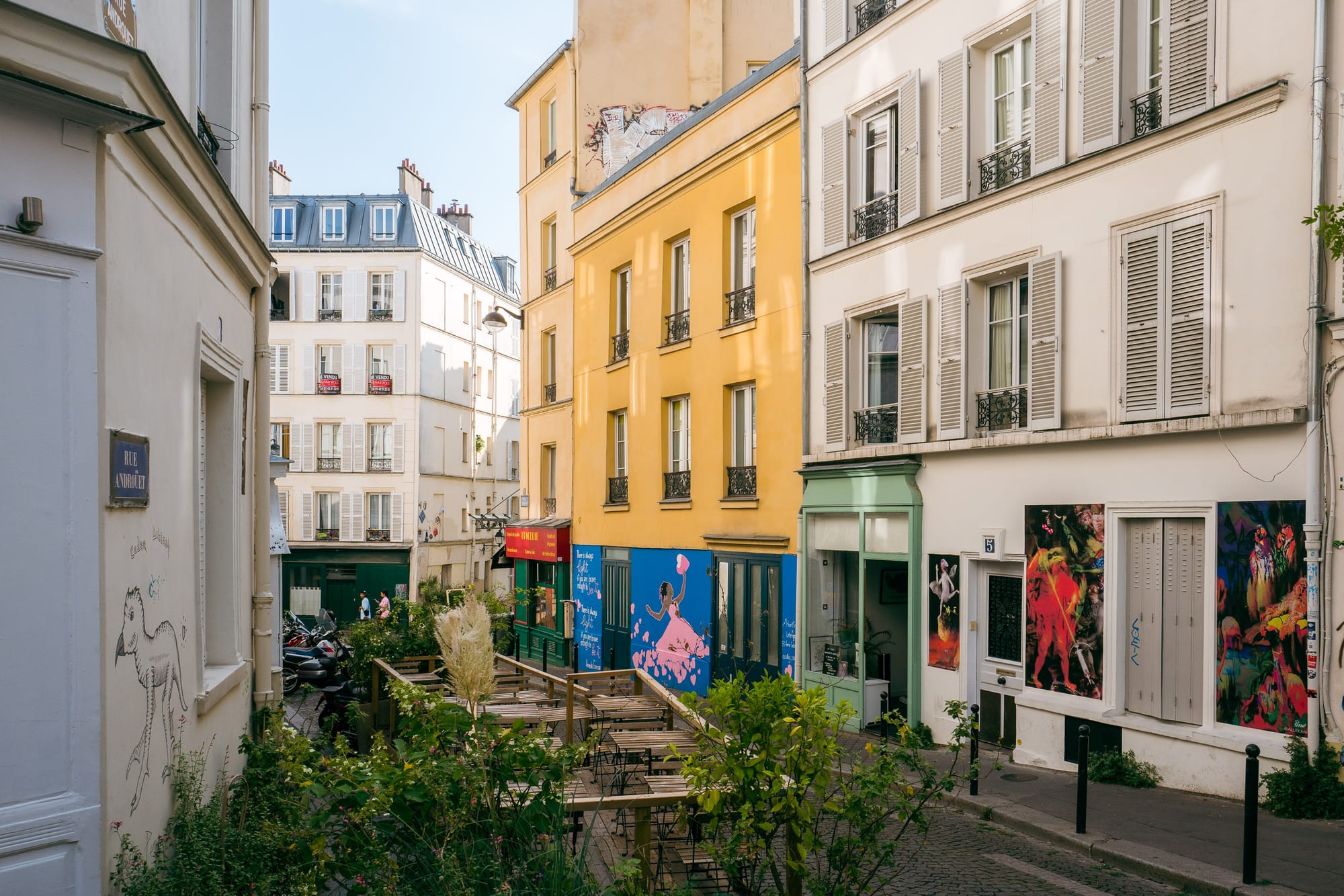 A Parisian street without people. Most of the buildings are white/cream, but one is yellow with a blue ground floor.Sunlight reflects off windows and onto other buildings. There are small green trees and it is sunny.