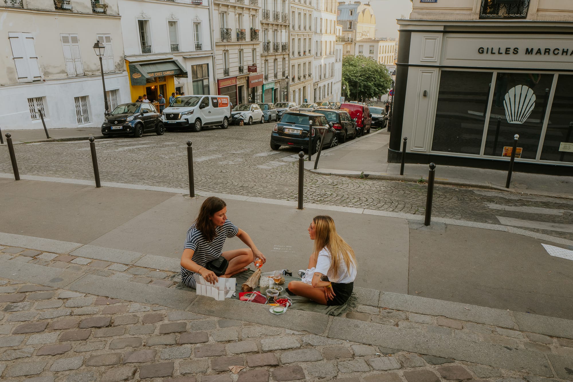 Two women have a picnic on the pavement. They have wine, bread, and fruit. Behind them is a road leading down a hill with shops and cars parked outside.