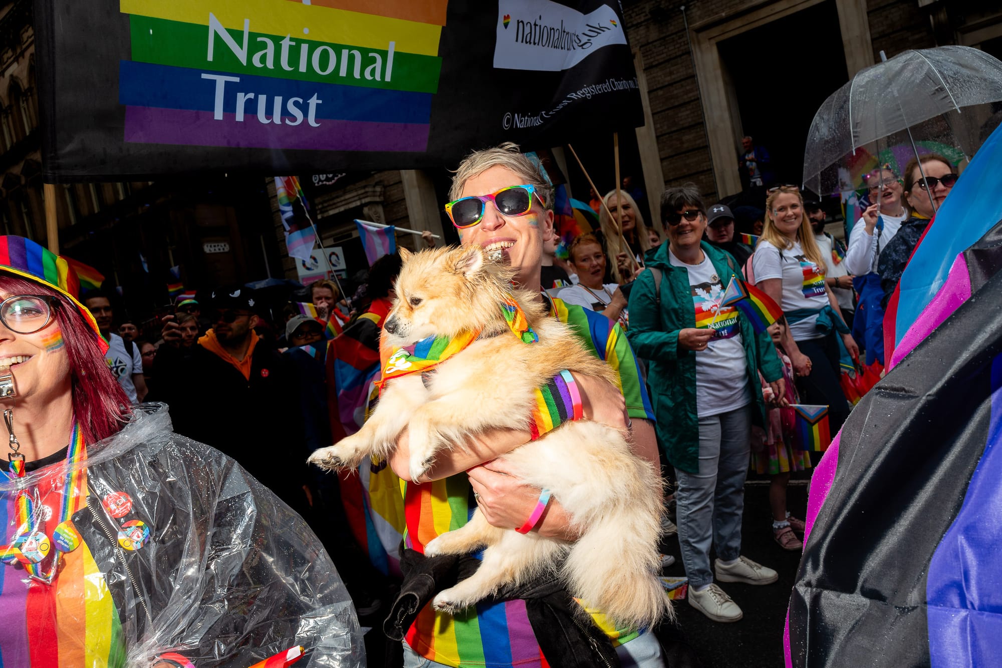 A white person holds up their dog during a Pride March. The dog has a rainbow neck tie on.
