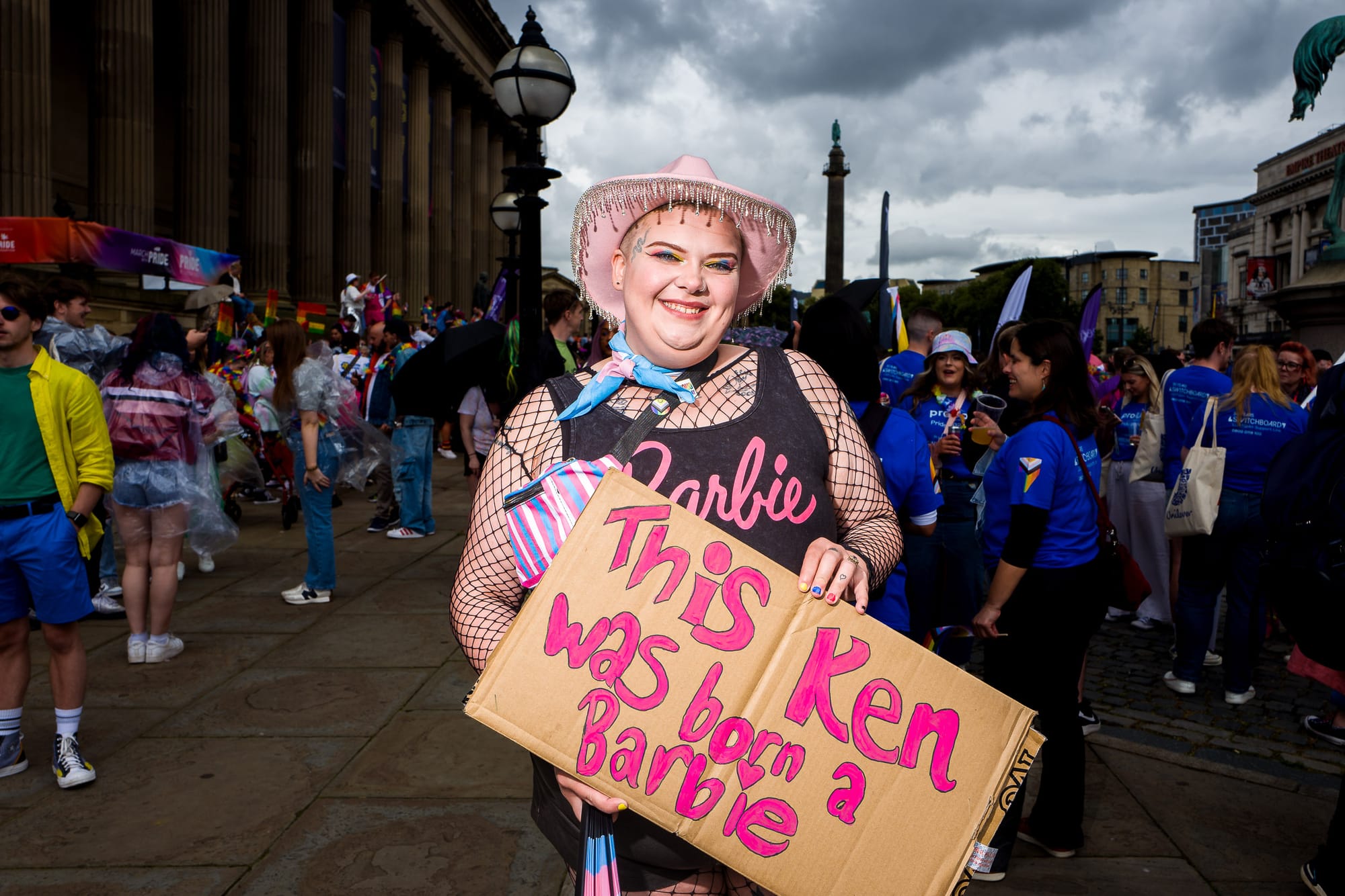 A trans man in a fishnet body suit and Barbie top holds up a sign saying This Ken was born a Barbie.