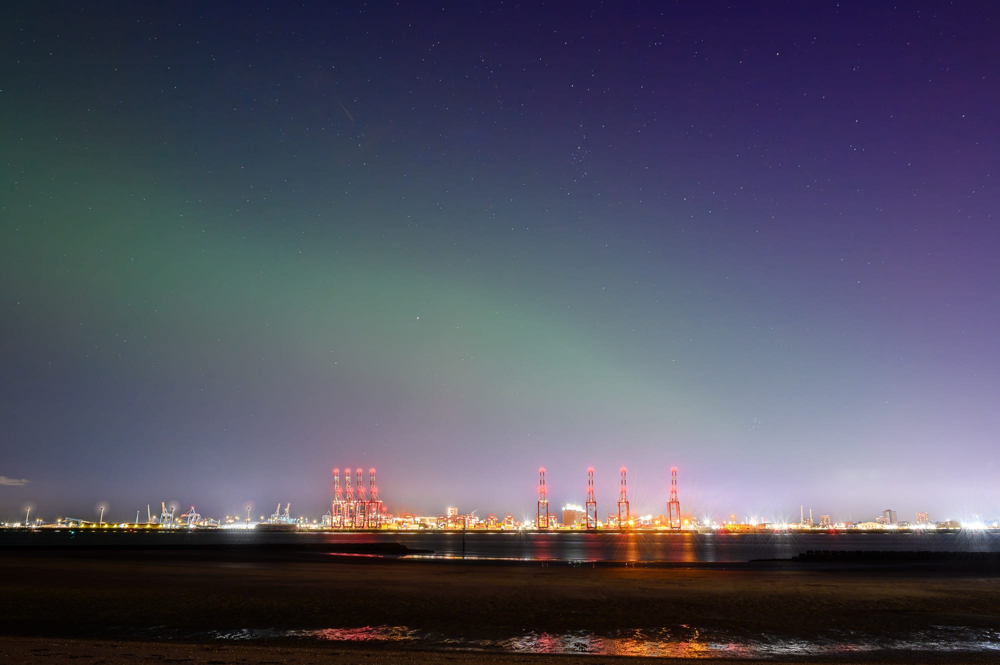 The nights sky is blue and purple from the aurora. Above the red cranes of the docks is a green whispy aurora. The tide is out.