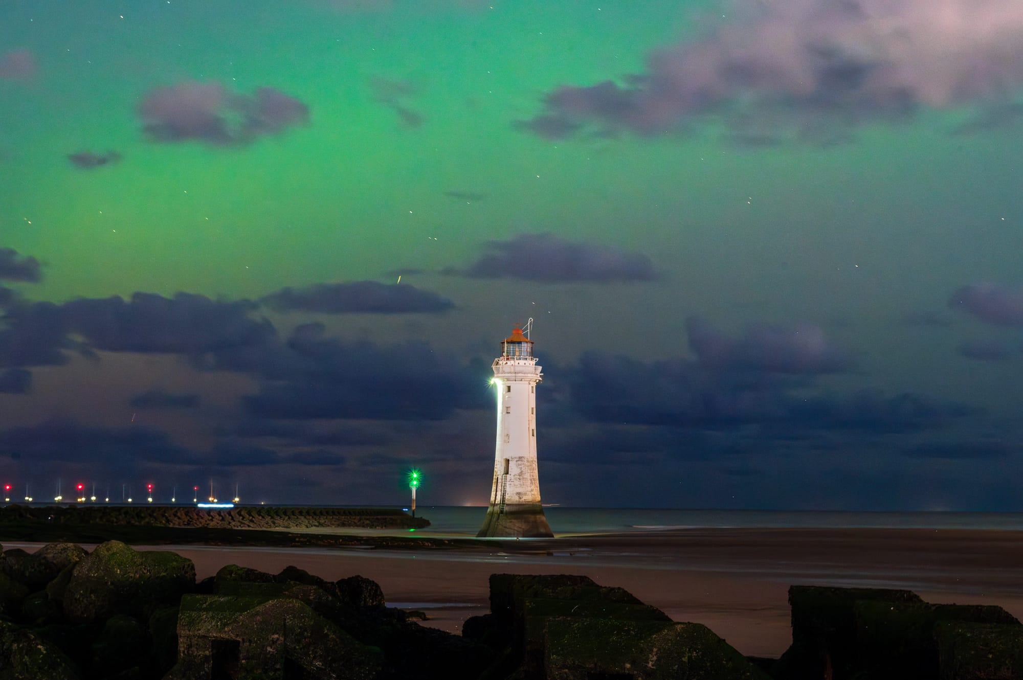 The tide is out around a lighthouse. The sky is green from the aurora. There are sea defence rocks around the lighthouse.