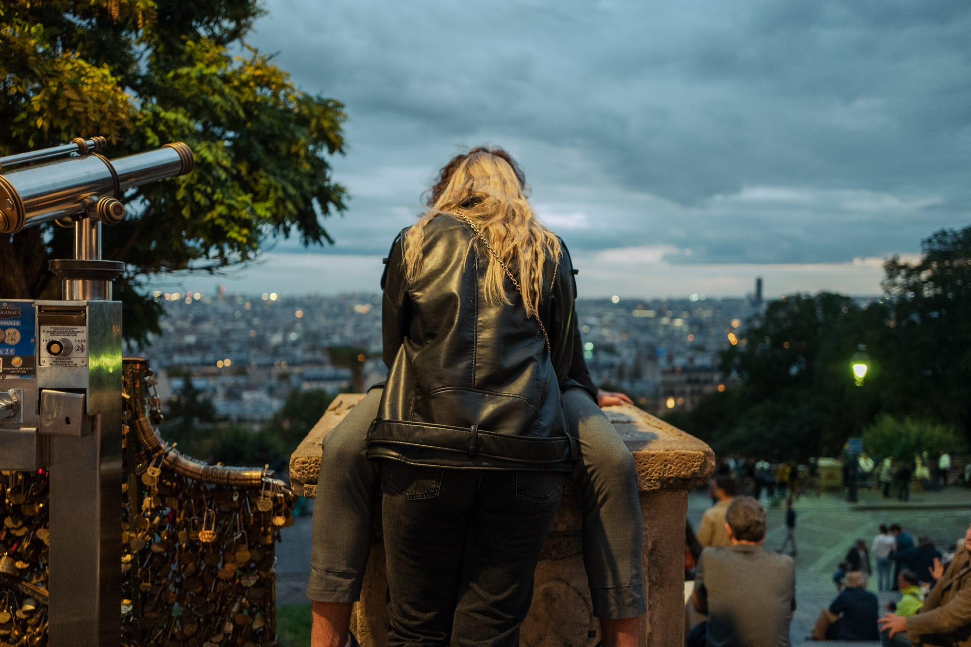 On the steps of Sacre-Coeur, overlooking the skyline of Paris, a person sits on top of a small concrete platform. Another person stands in front of them a leans in for a kiss.
