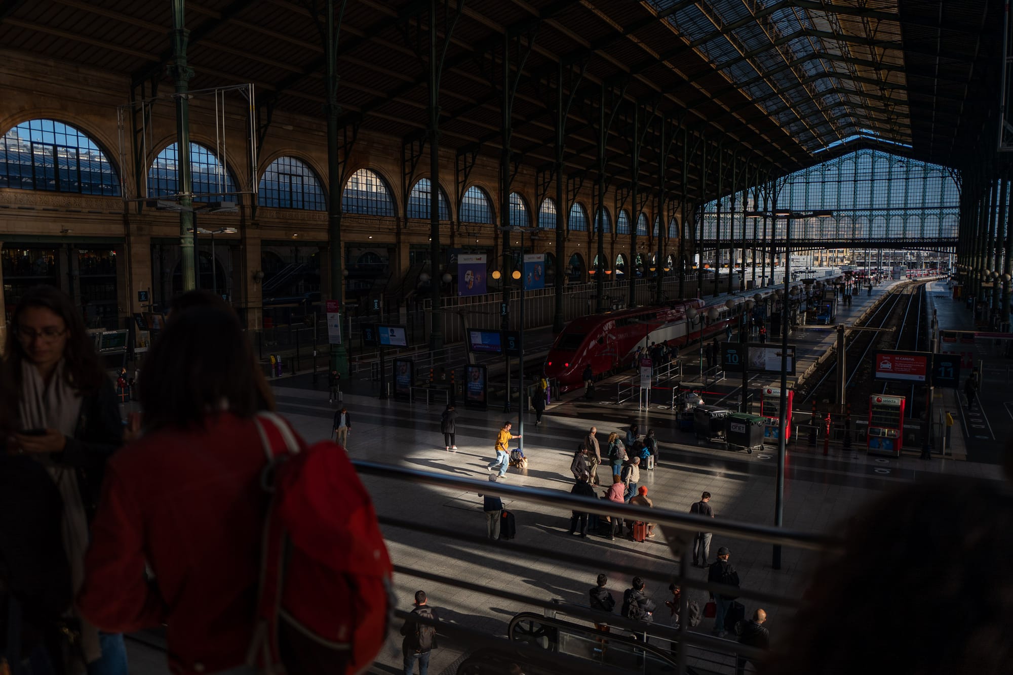 In a large train station sunlight creates a spotlight effect on a single person. They are wearing an orange top and jeans, and they are dancing alone in the spotlight.