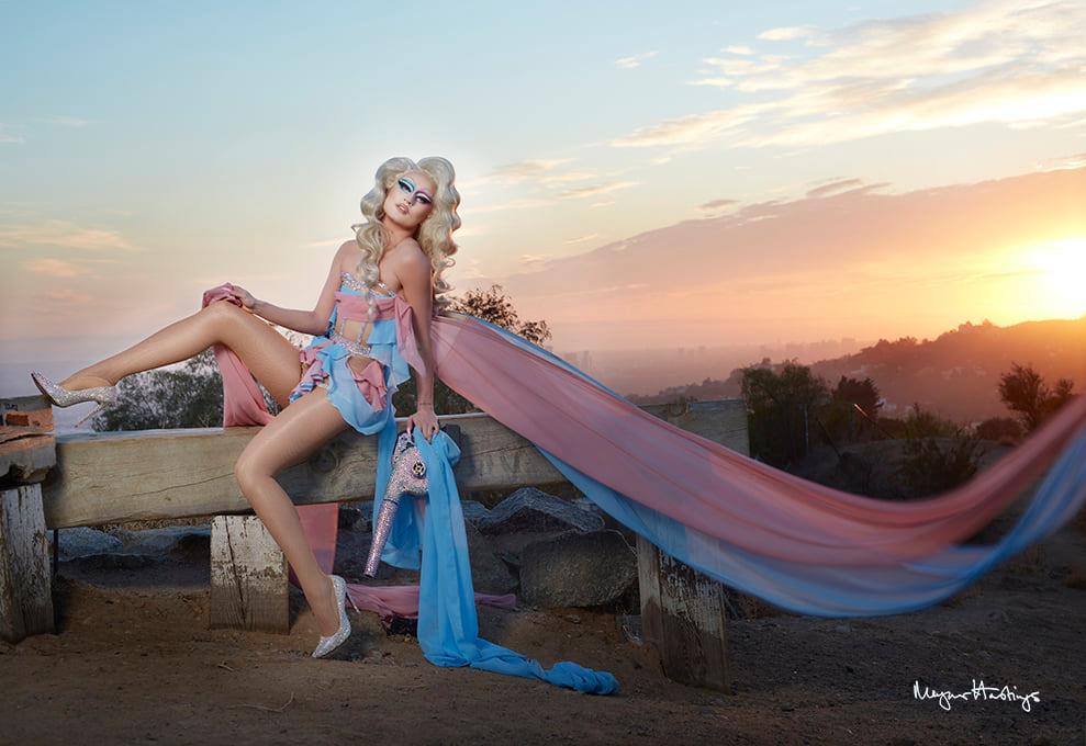 A drag queen wears a trans-themed dress with a very long trial flying in the wind at sunset.