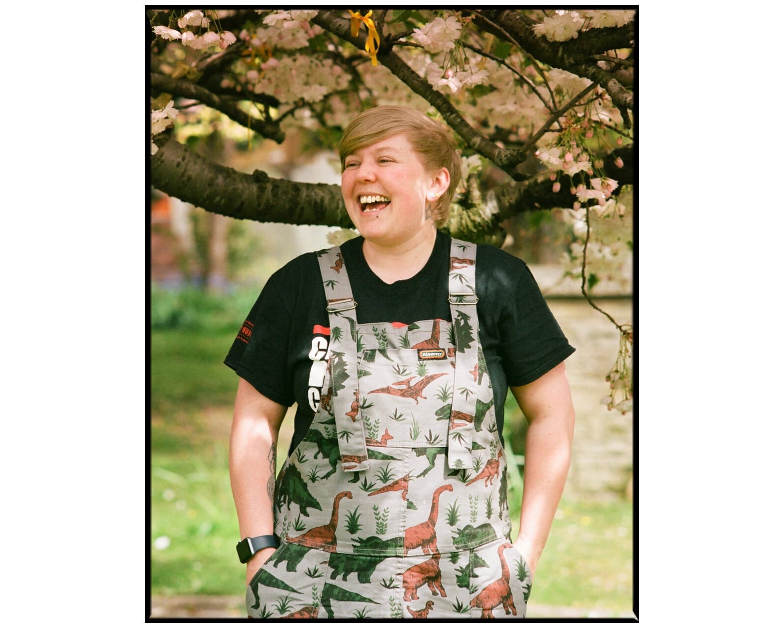 A woman stands by a tree, laughing, while wearing dinosaur dungarees.