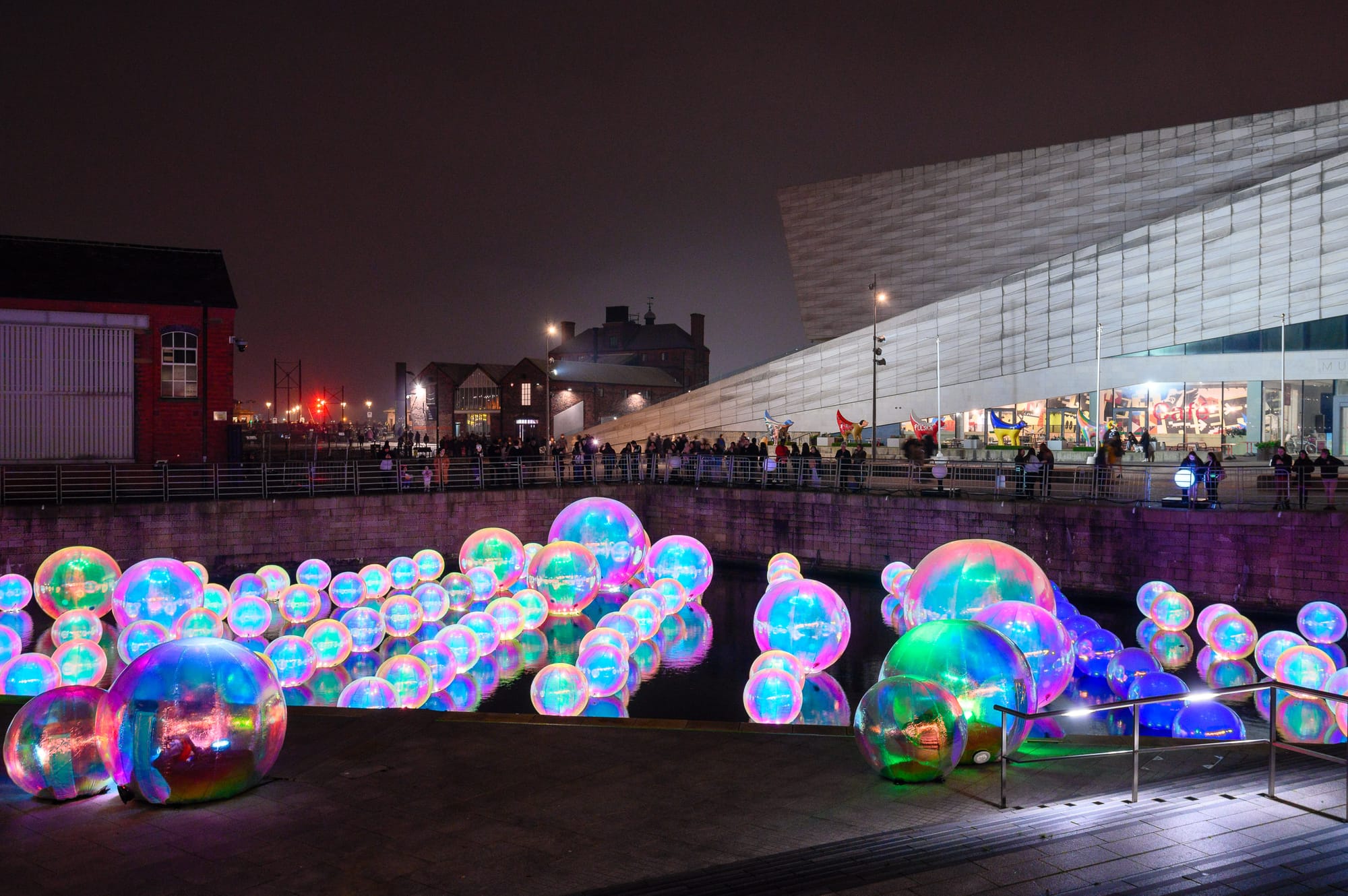 In the canal near the Museum of Liverpool are giant inflatable balloons glowing in various pastel colours. People look from above.