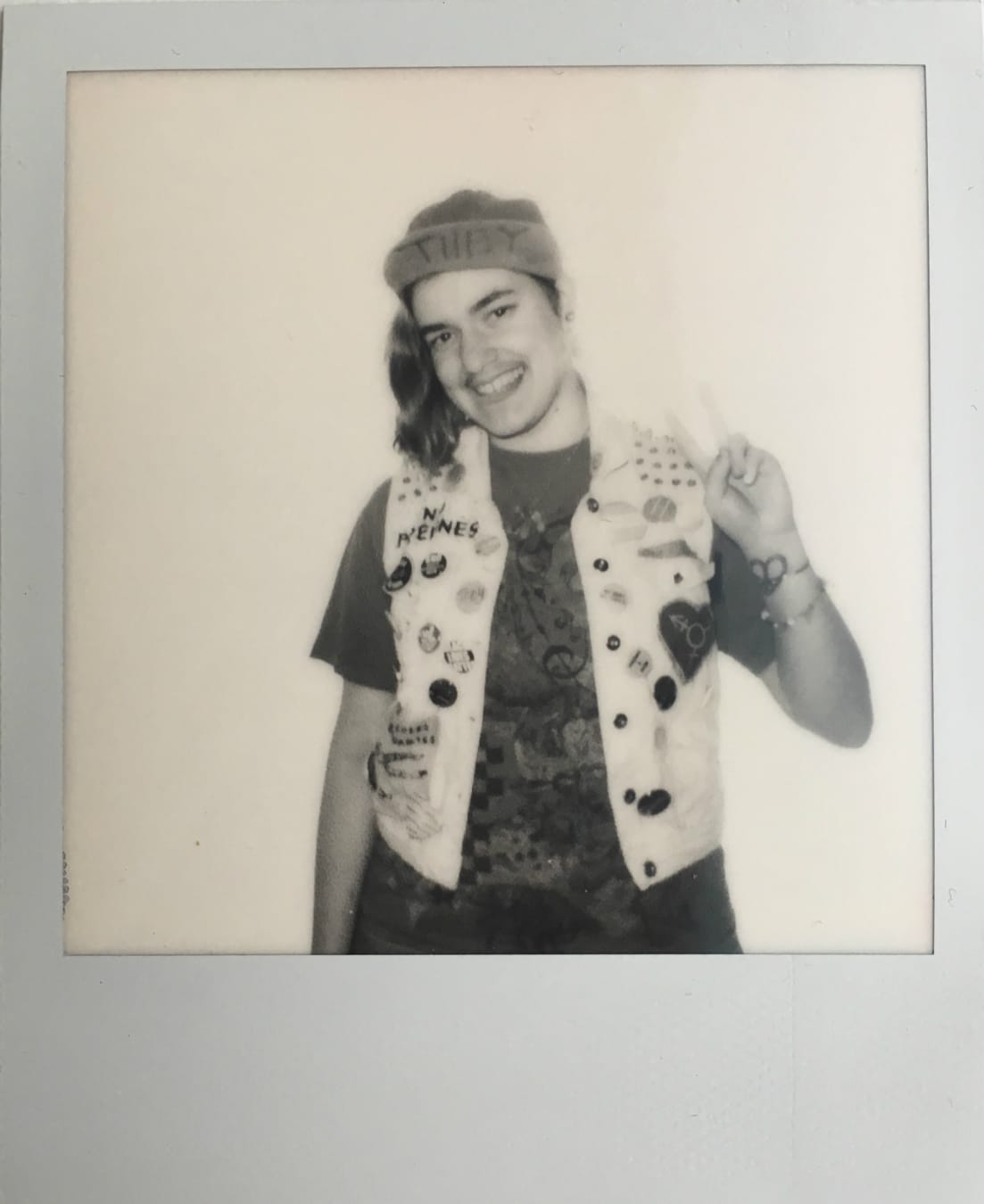 A polaroid of a person holding up a peace sign to the camera. They have a beanie on with the word 'they' on it.