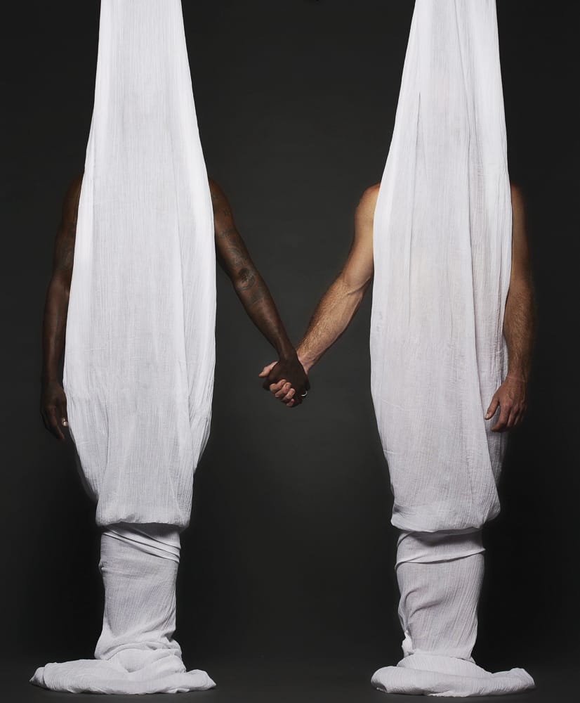 A black man and a white man stand underneath sheets and hold hands.