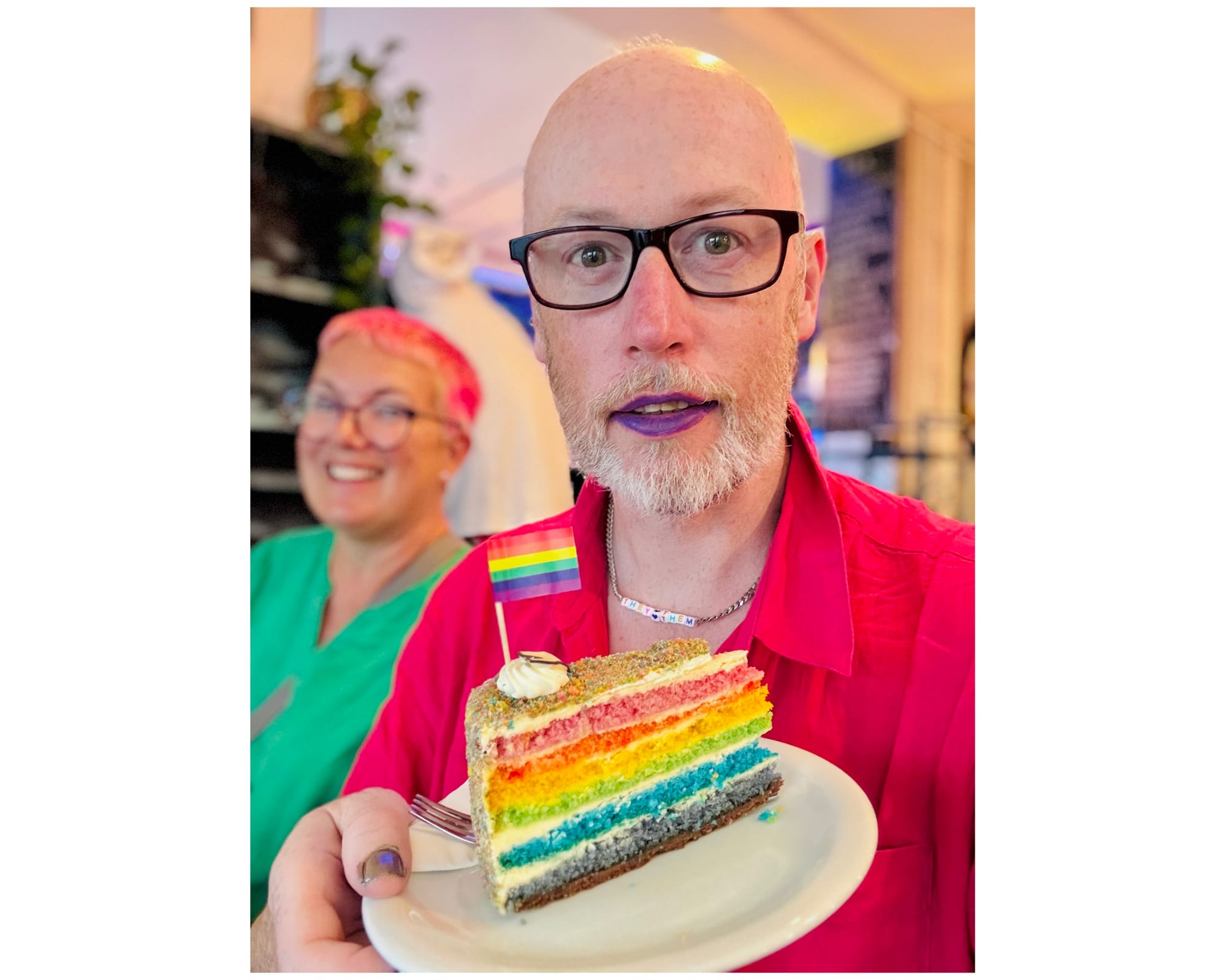 Non-binary person holding up a rainbow cake with a rainbow flag on. They are wearing a they/them choker, with purple lipstick and glasses. Behind them their wife smiles.