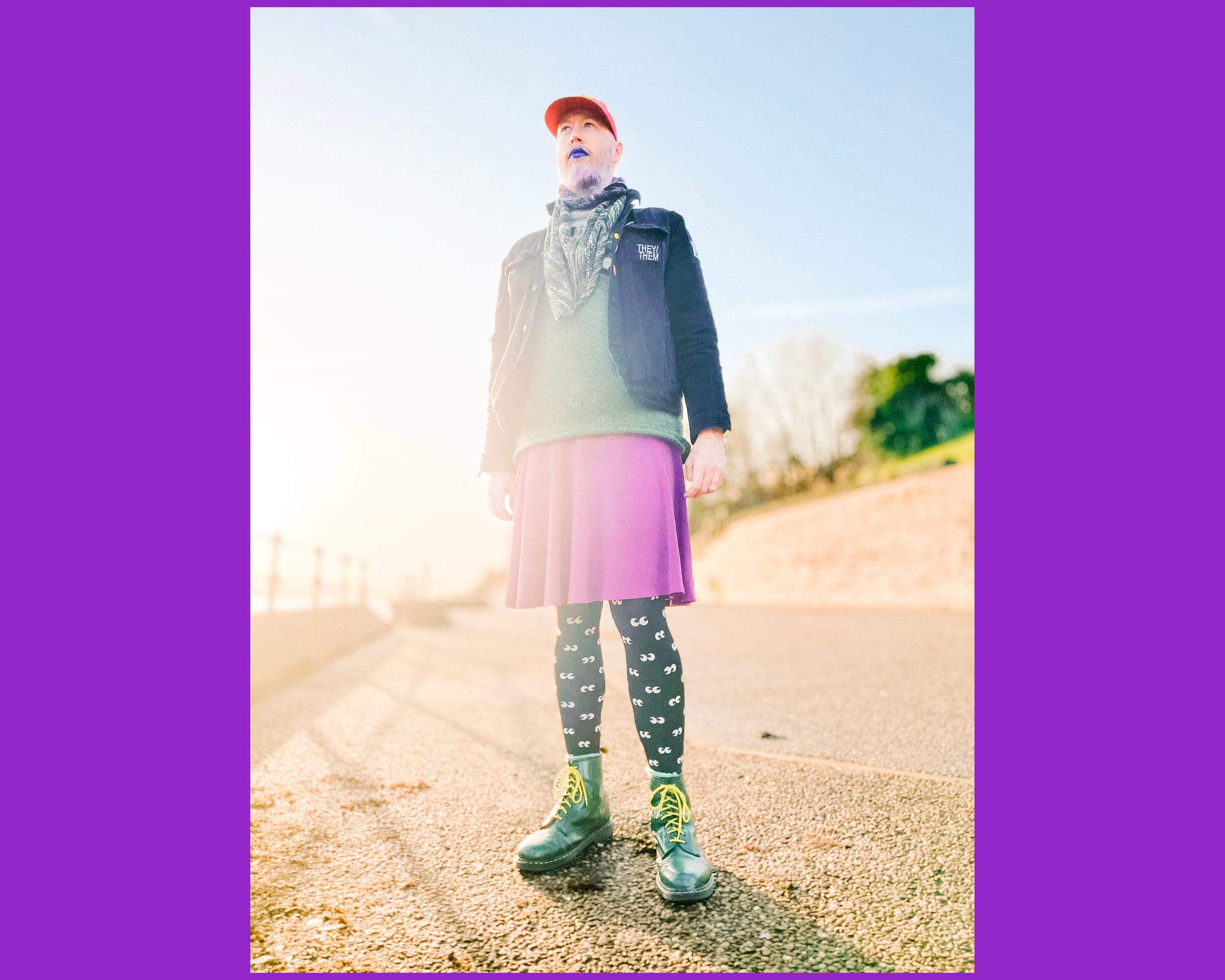 A non-binary person wearing a purple skirt, black tights with eyes on, green boots, a denim jacket with they/them on and makeup looks to the distance.