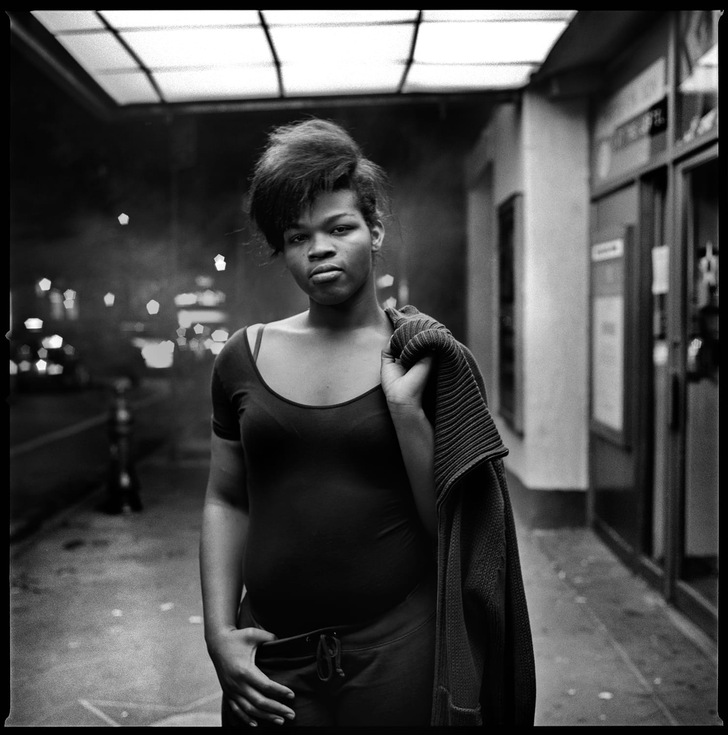 A black woman with big hair looks to camera and holds their coat over their shoulder.