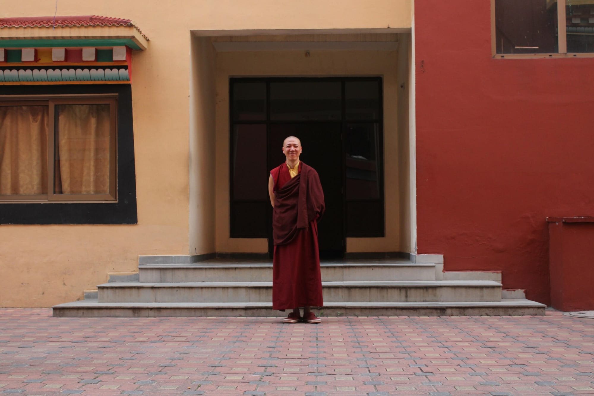 A monk wearing red and yellow robes stands outside a red and yellow building.