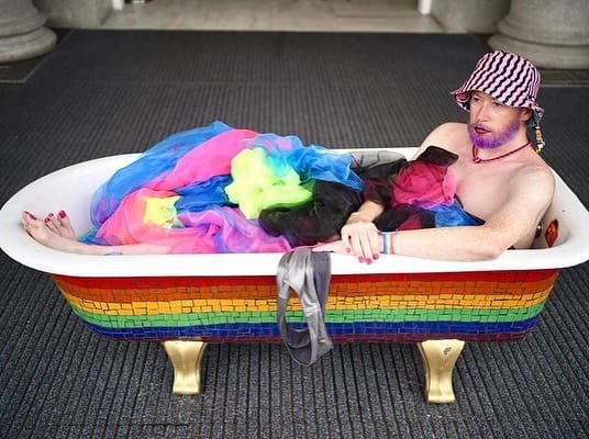 Apparently naked non-binary person in a rainbow bath tub covered by rainbow cloth