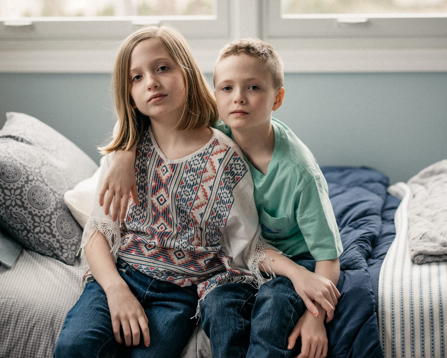 Two twins of different genders sit next to each other. One has their arm around the other, and the other reaches their arm across their siblings leg to rest on their arm.