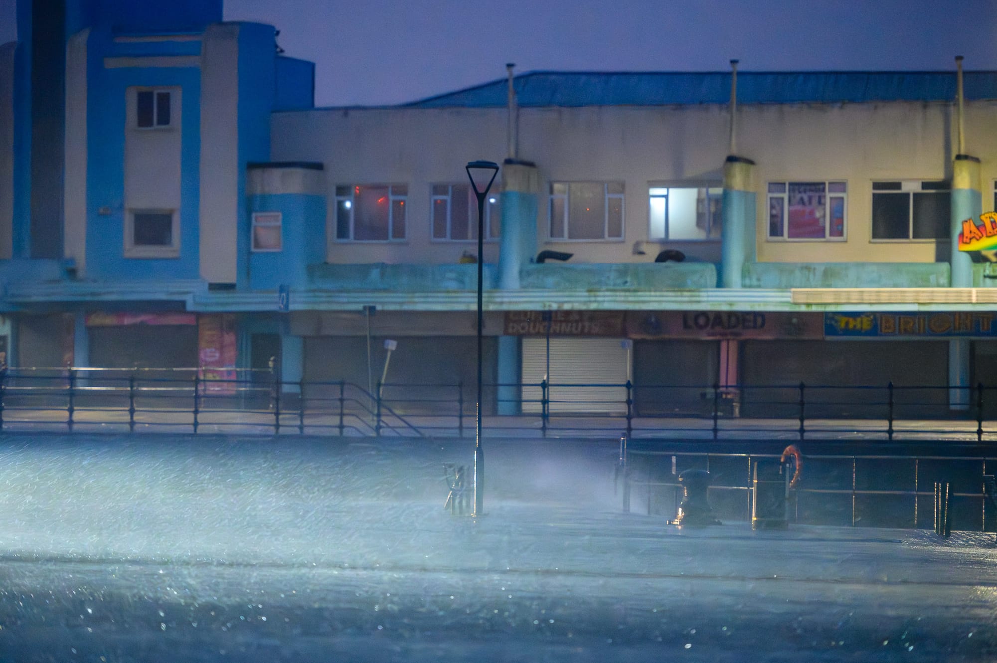 The wind is lifting water out of a lake and across an empty car park at dawn.