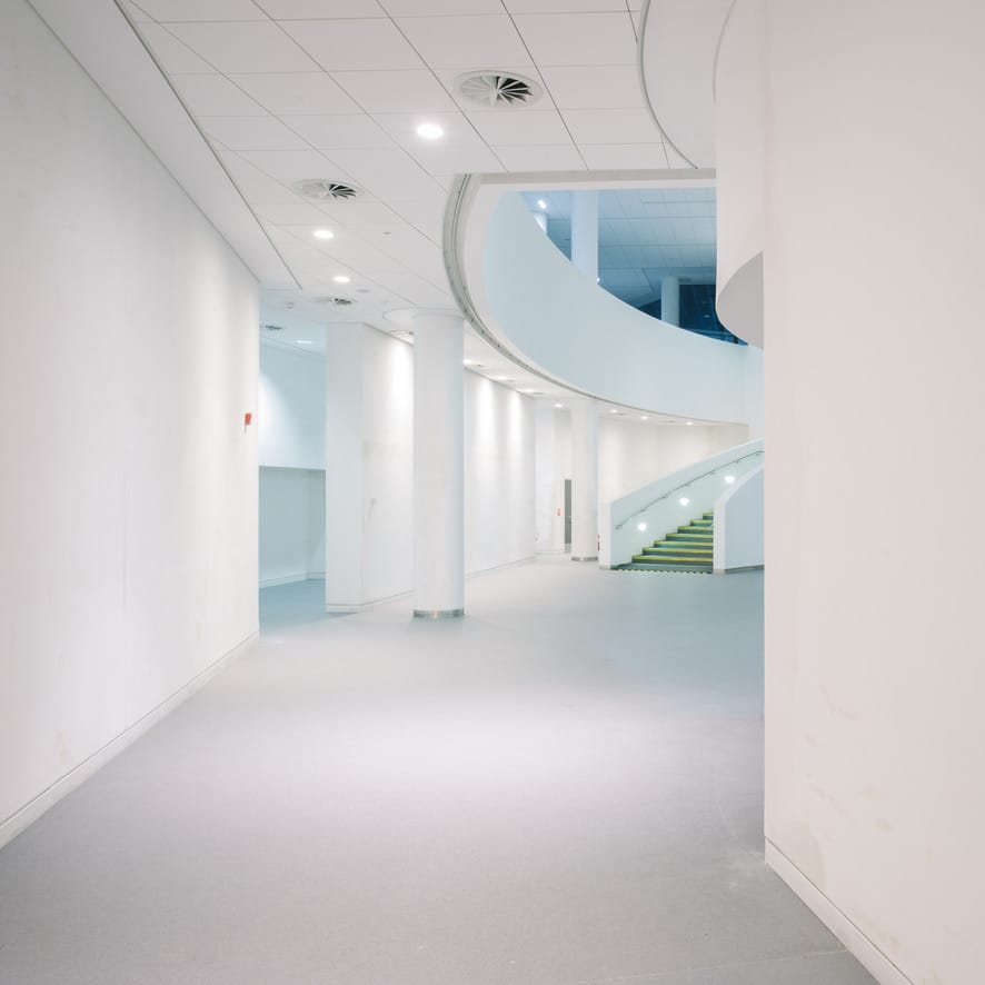 A spacious, modern interior with a smooth, light-colored floor and plain white walls. A curved staircase can be seen in the background, leading to an upper level. The area is well-lit with ceiling lights and has a minimalist design, creating a clean and airy atmosphere. There are no visible furnishings or decorations, emphasizing the simplicity of the space.