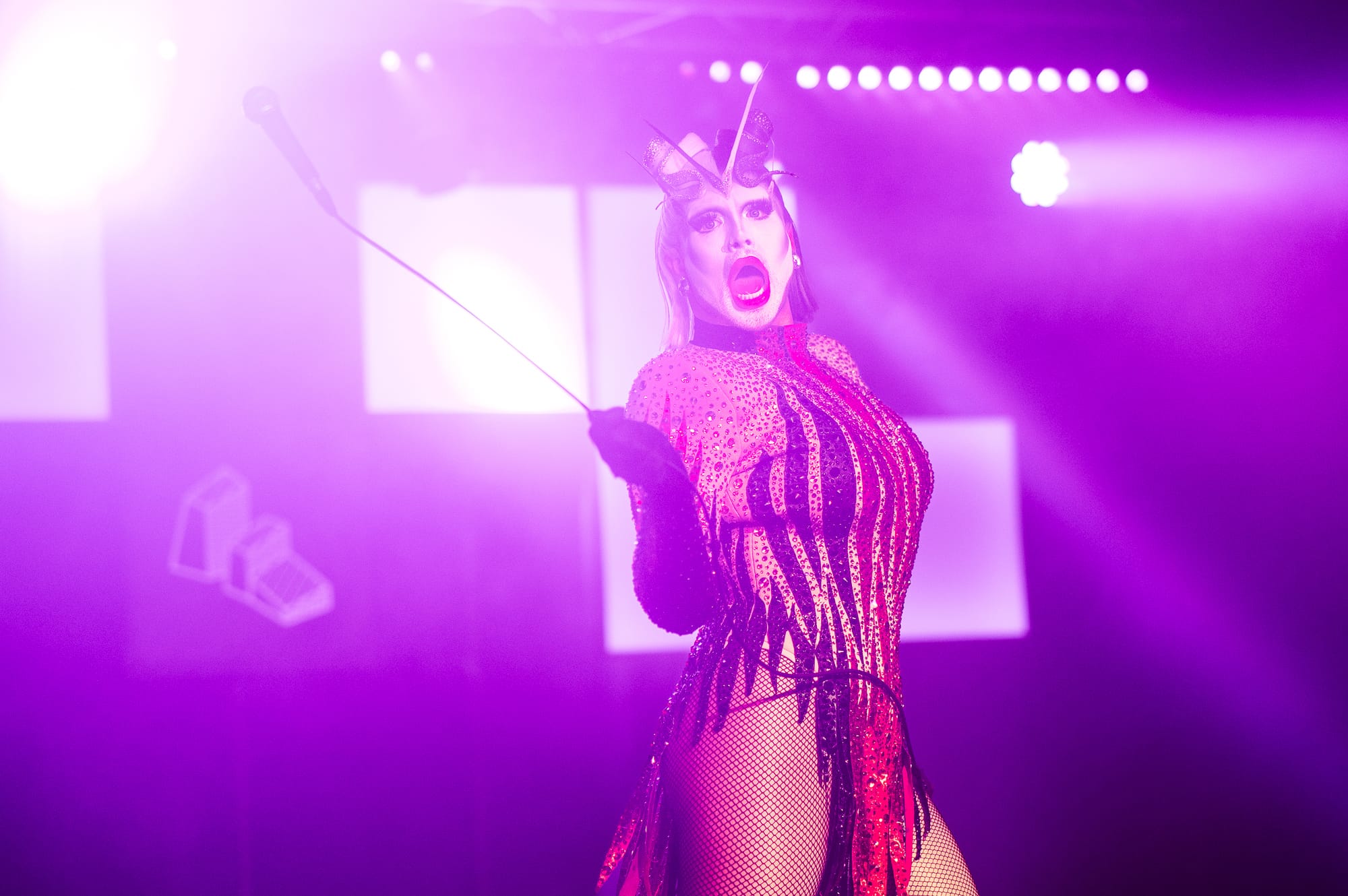 A performer in a sparkling, form-fitting outfit with black gloves is dramatically posed on stage, holding a whip, with intense lighting creating a vibrant purple atmosphere.