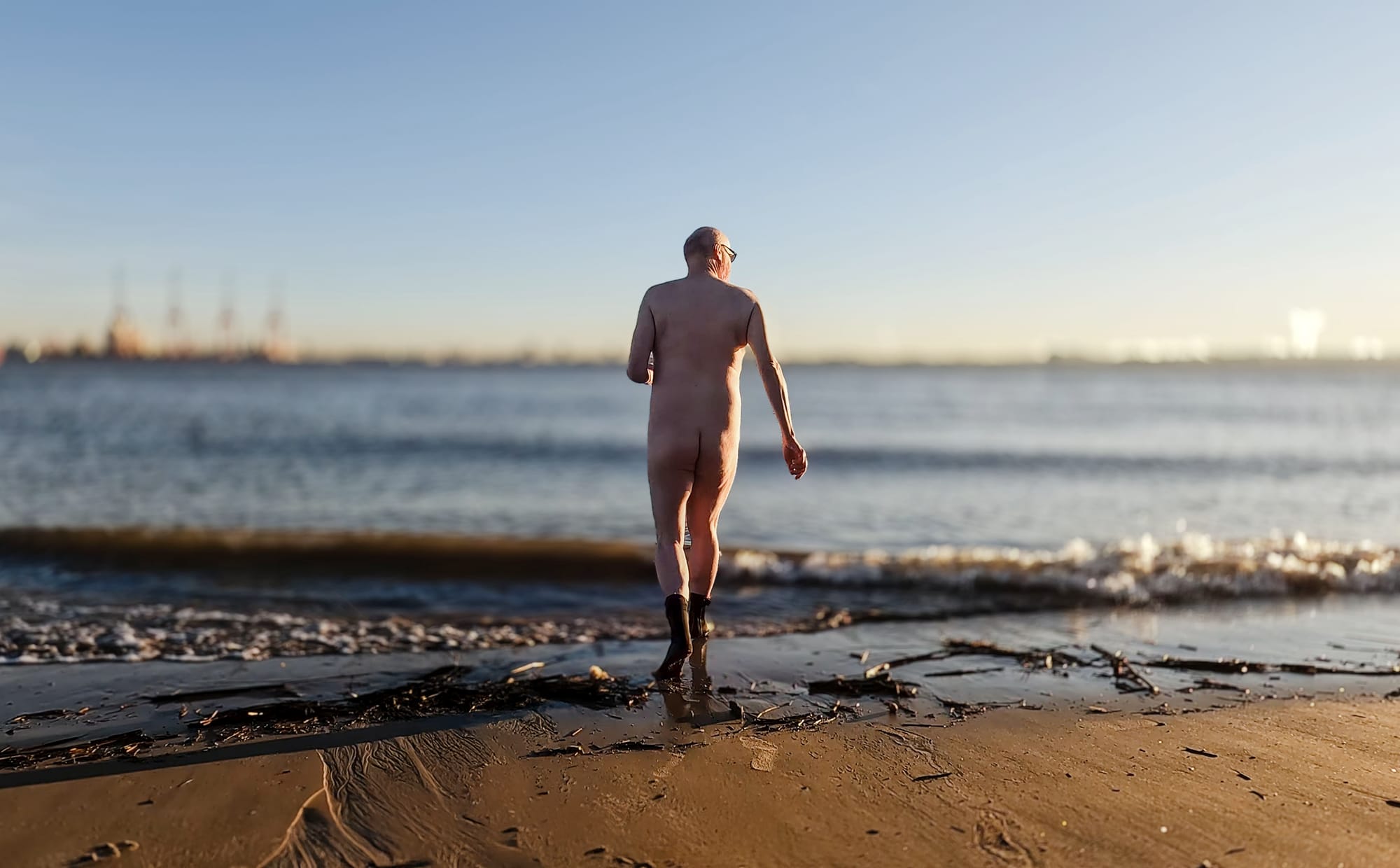 A person walks away from the camera along the shoreline at sunset, with the ocean waves gently lapping at their feet. They are nude and wearing only black boots. The scene is peaceful, and the sun casts a warm glow over the water and sand, while distant industrial structures are visible on the horizon.