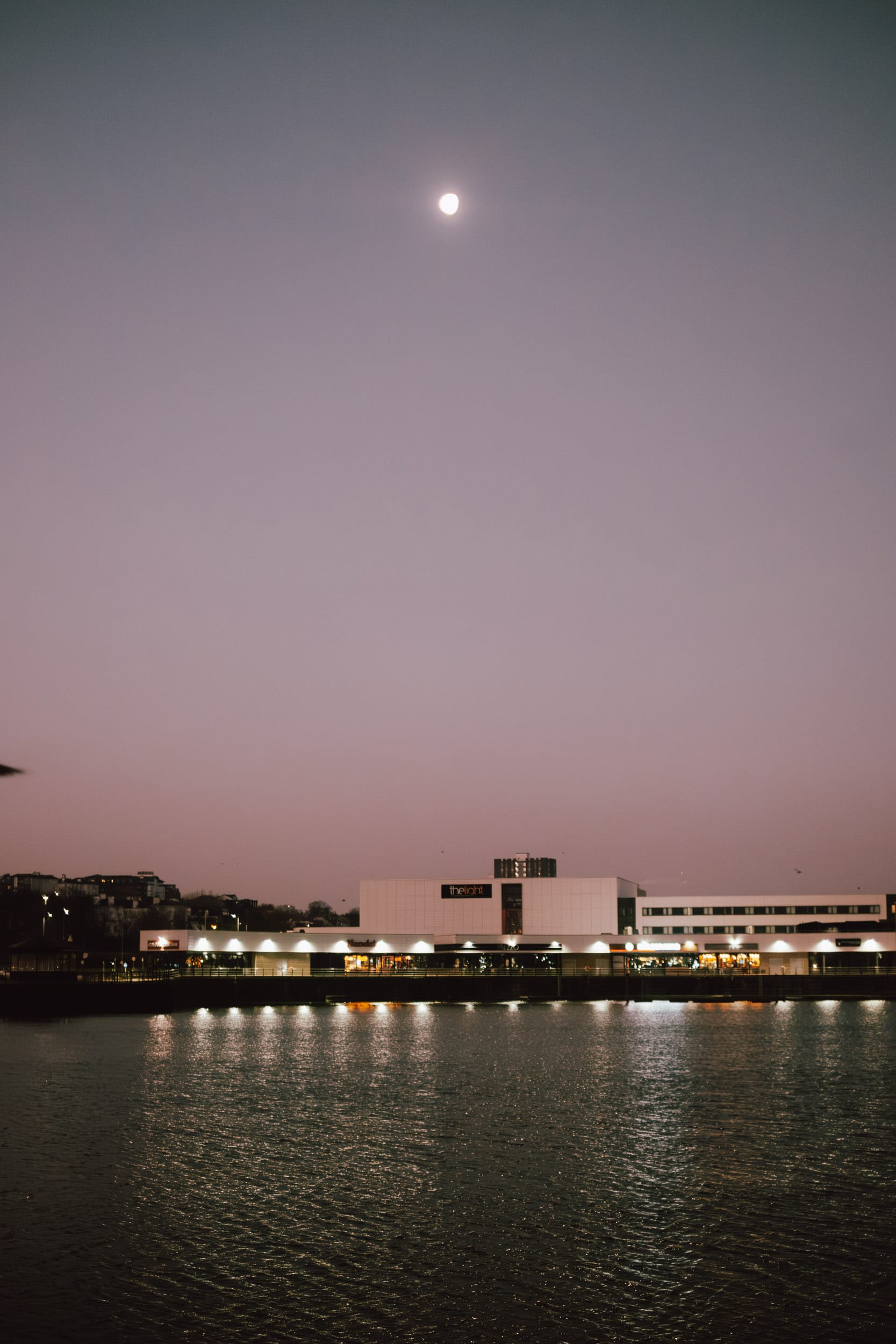 A riverside view with a building reflecting on the water, under a sky with the moon visible. The building appears to have lighted sections along the water's edge, with other buildings in the background.