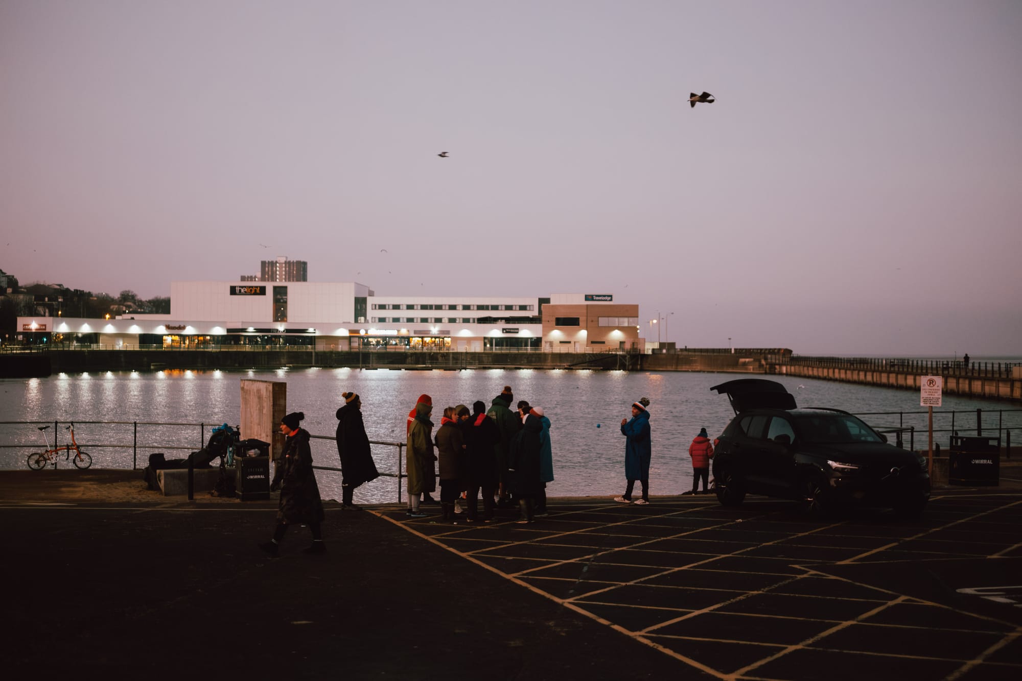 A group of people gathered near a waterfront at dusk, with a modern building in the background that has signs indicating businesses. A bicycle is parked nearby, and a car is parked in a designated area. The scene includes a calm body of water reflecting lights from the building, and there are a few people standing away from the group.