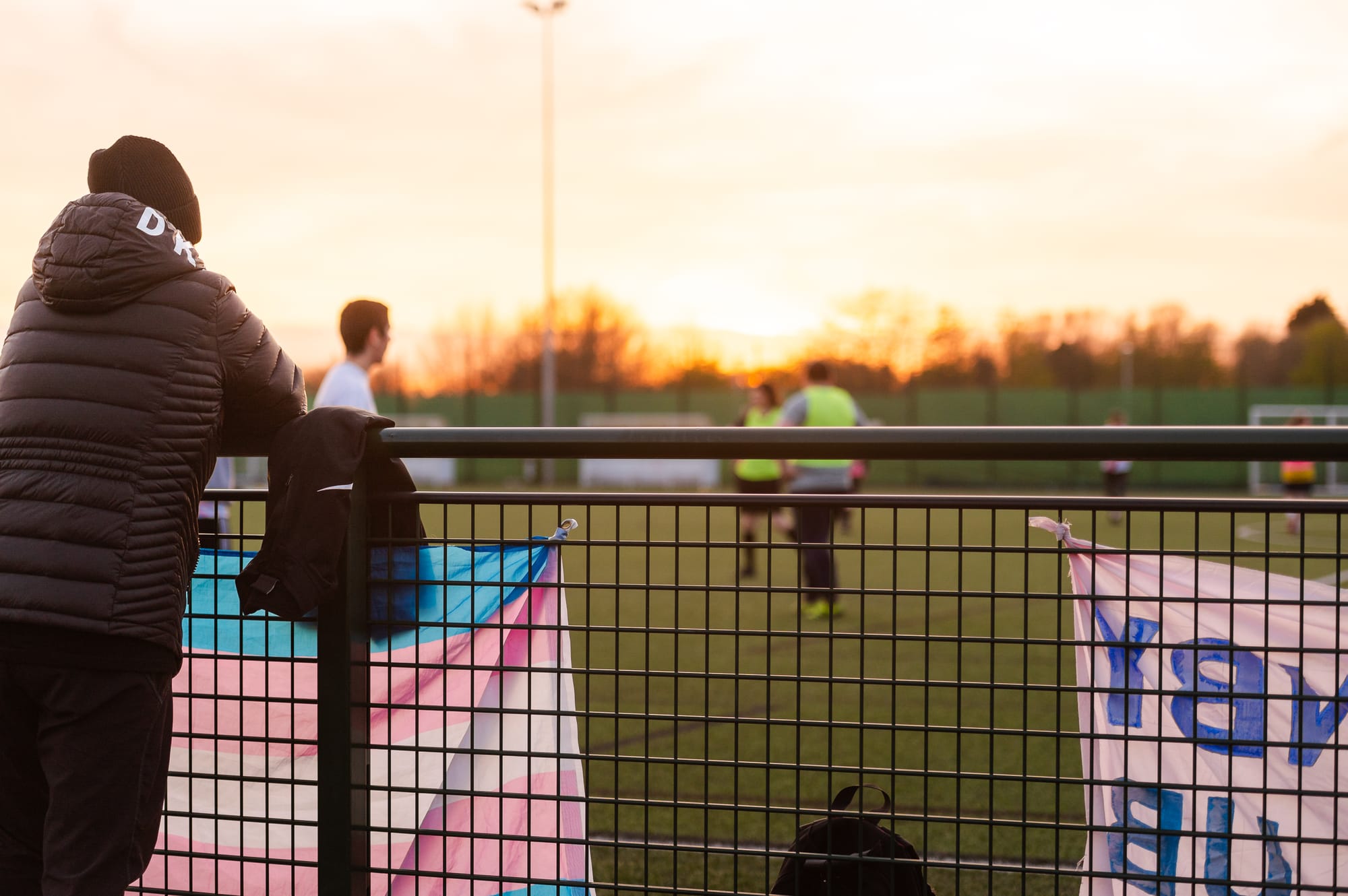 A person wearing a black puffer jacket and a black beanie stands with their back to the camera, leaning on a fence at a soccer field. A trans flag and another flag are attached to the fence. Several people are playing soccer in the background, the setting sun casts a warm glow over the scene.
