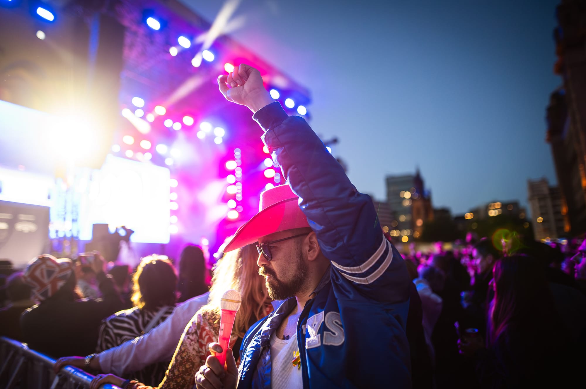 A man in a blue jacket and pink cowboy hat raises his fist in the air at an outdoor concert at night. The stage is brightly lit with pink and blue lights, and the crowd is visible in the background.