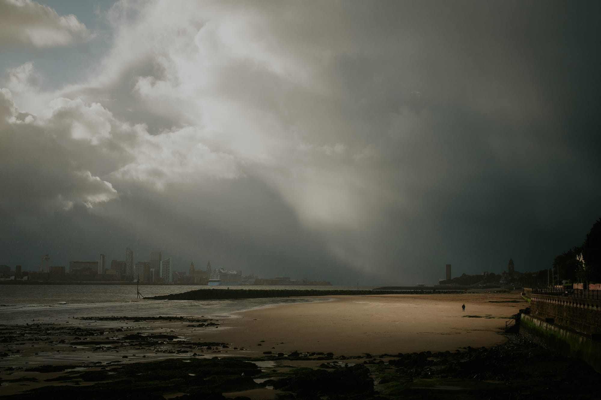 A lone figure lit by dramatic light standing on a beach at low tide.