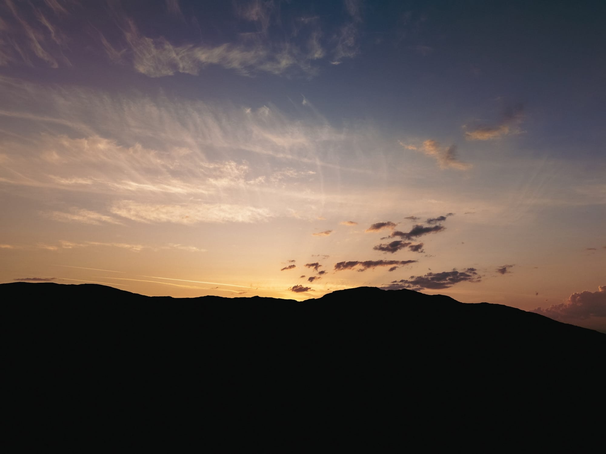 A beautiful sunset scene featuring a gradient sky transitioning from deep orange and yellow at the horizon to a soft purplish-blue above, with wispy clouds scattered across the sky. The silhouette of hills is visible in the foreground, creating a serene and tranquil atmosphere.
