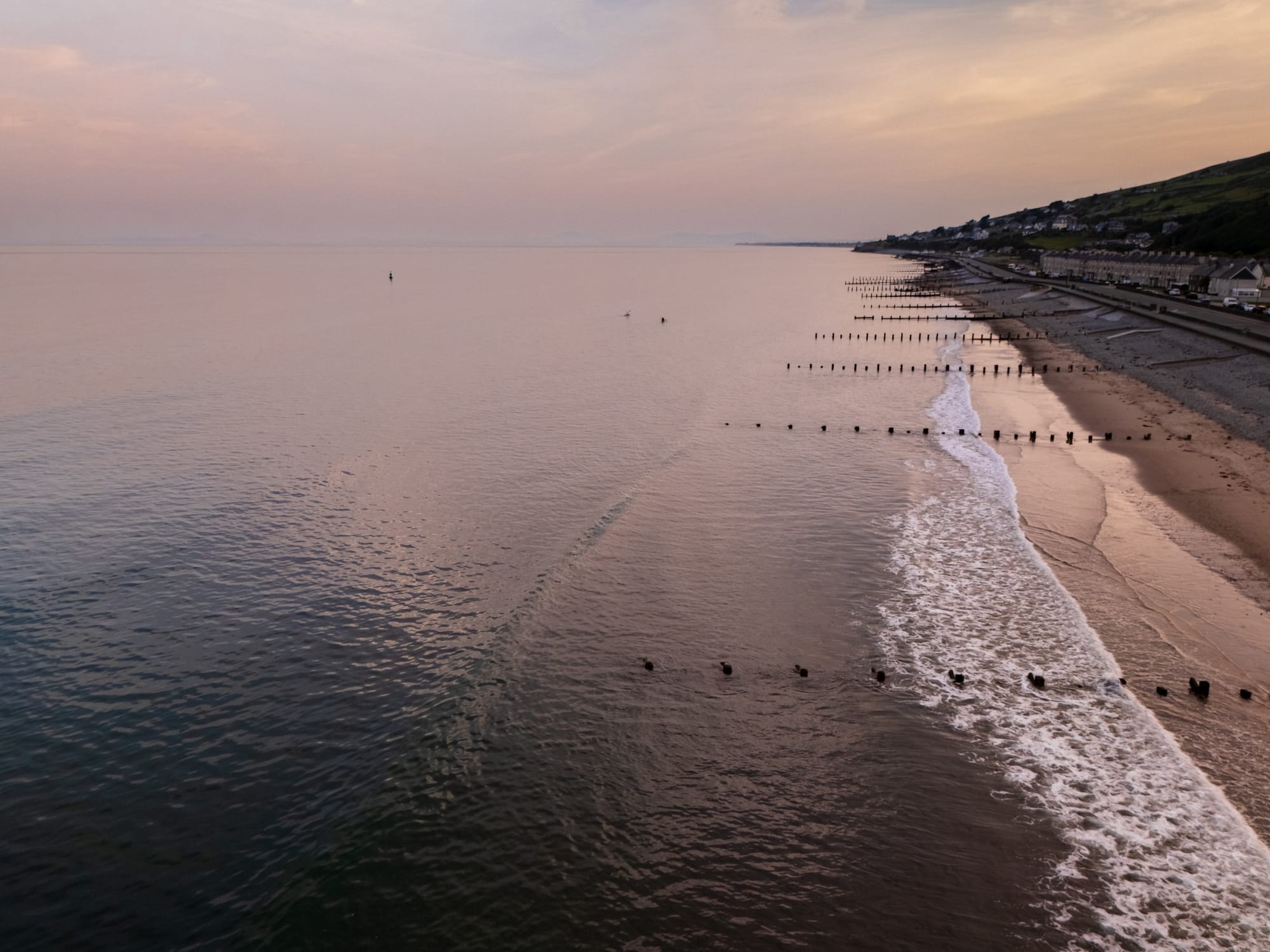 A serene coastal landscape at dusk, featuring a calm sea that reflects soft pastel colors in the sky. The shoreline is lined with gentle waves lapping against the sand, while wooden groynes extend into the water. In the distance, a small town is visible, nestled against the green hills. The overall mood is tranquil and picturesque, highlighting the beauty of nature at twilight.