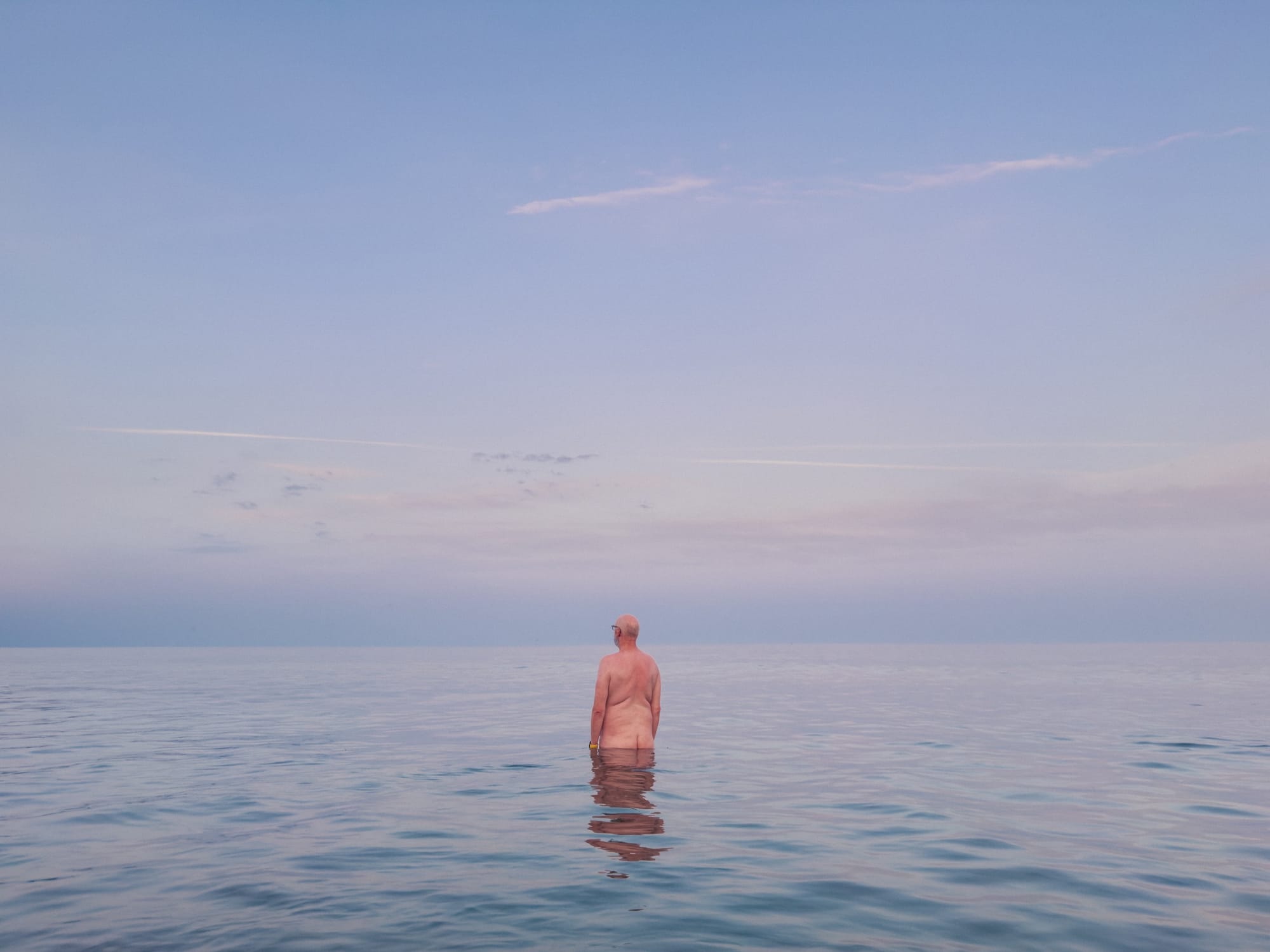 A person standing in calm water, facing away from the camera, with the horizon blending softly into a pastel sky at dusk. The water is still, reflecting the sky's hues, creating a serene and tranquil atmosphere.