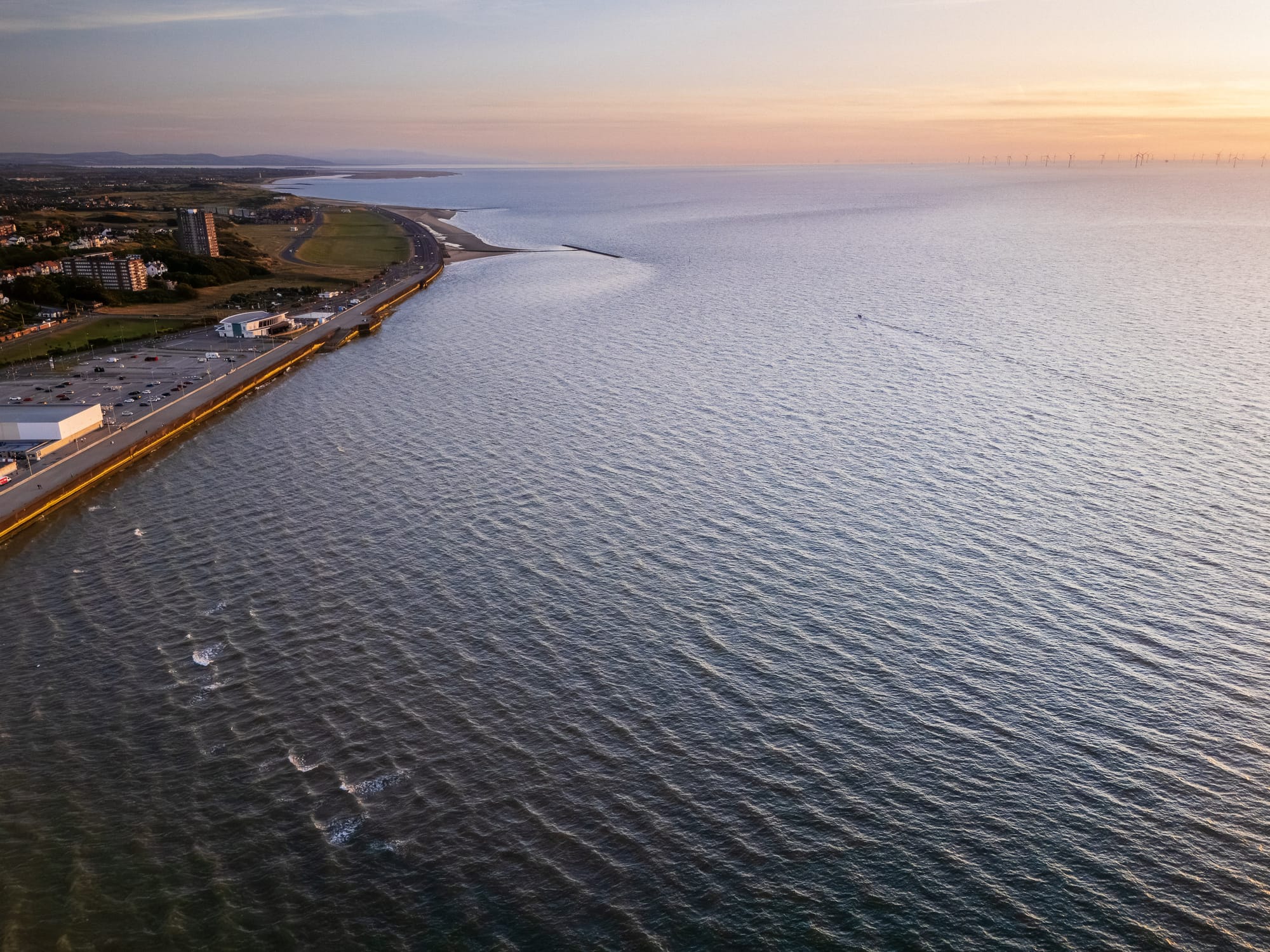 Aerial view of Liverpool Bay at sunset, showing the coastline, sea, and a few wind turbines in the distance.