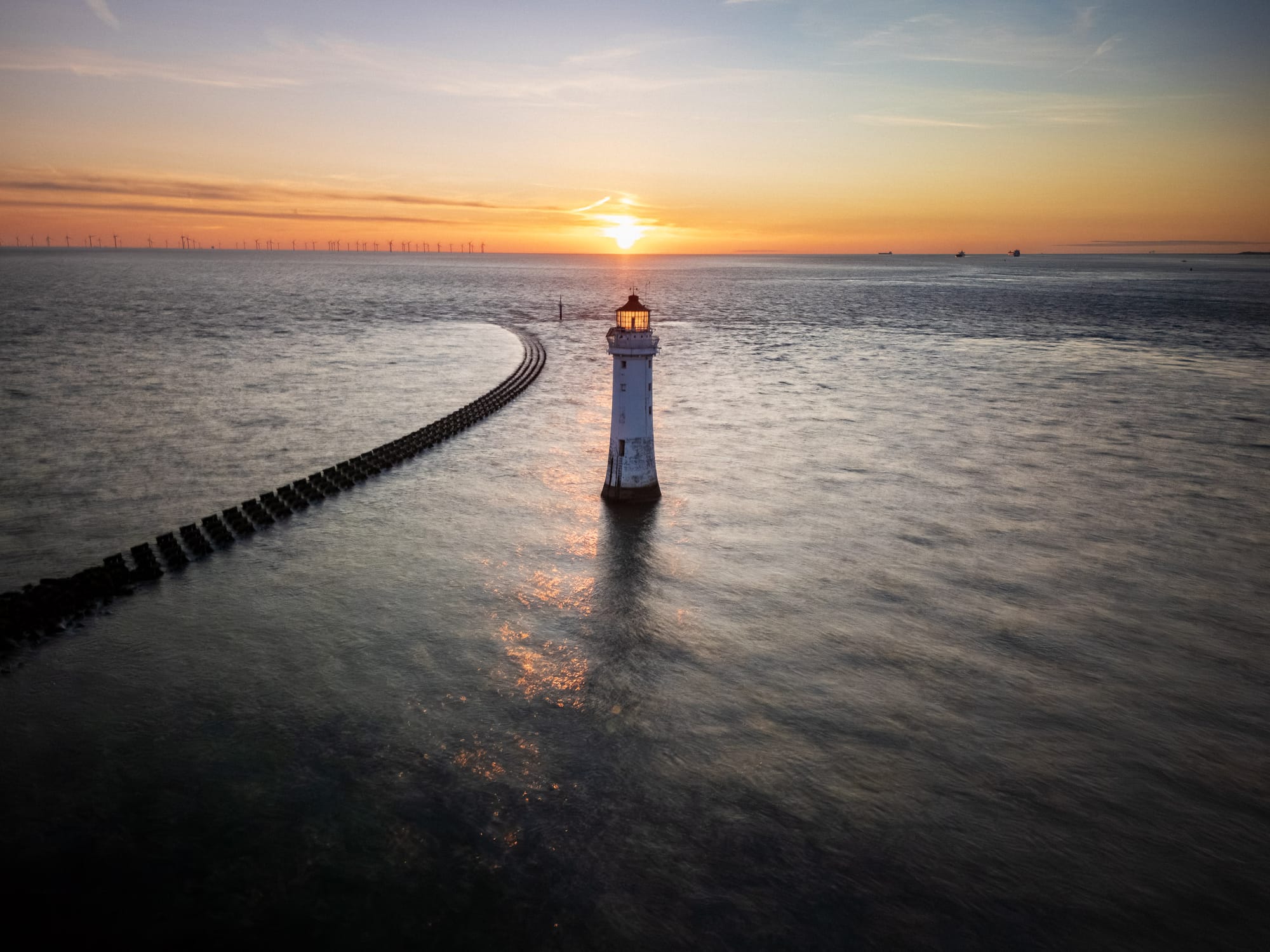 An aerial view of New Brighton Lighthouse at sunset, with a long breakwater extending into the sea.
