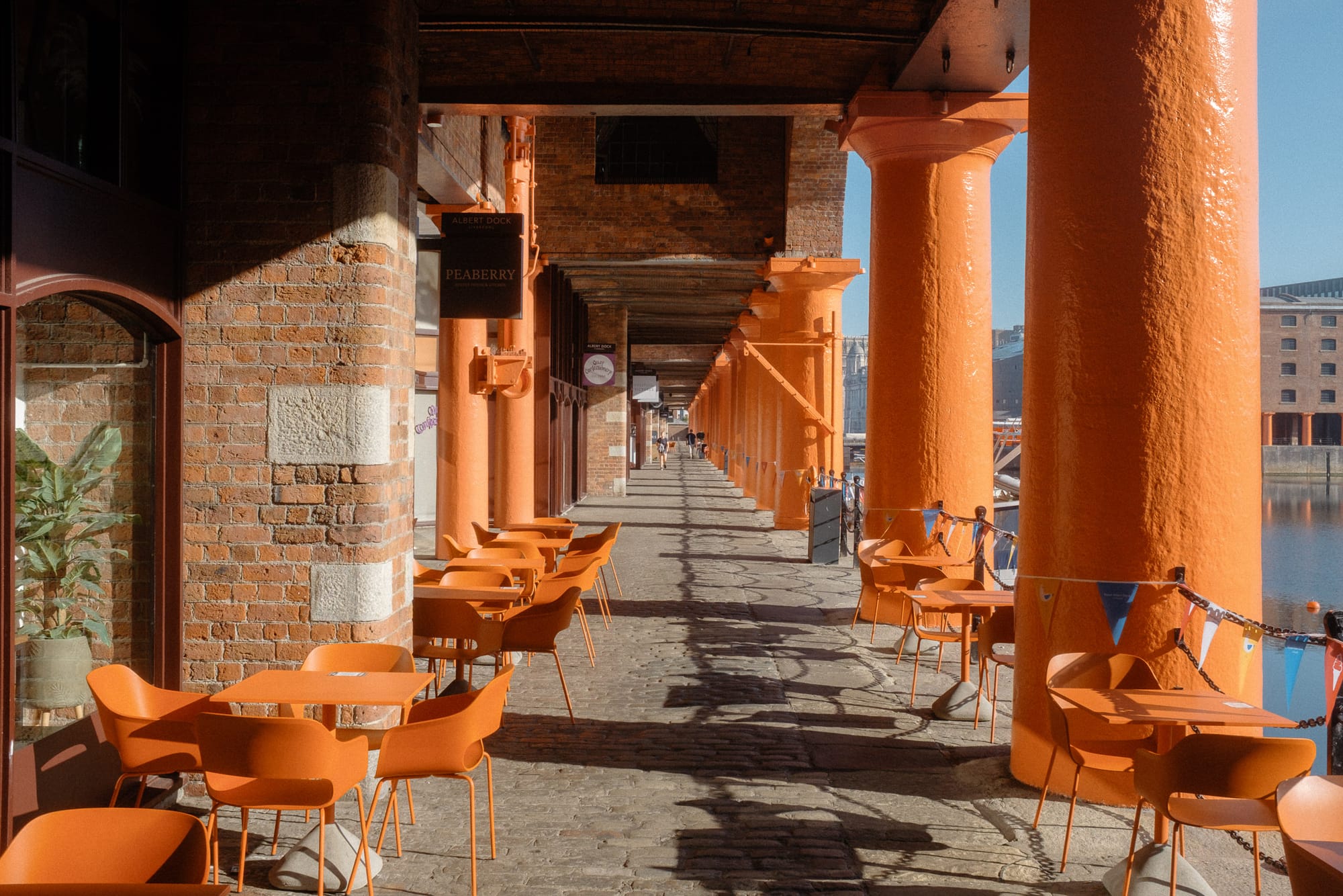 A sunny morning scene at Albert Dock in Liverpool, featuring orange pillars, outdoor seating, and a view of the water.