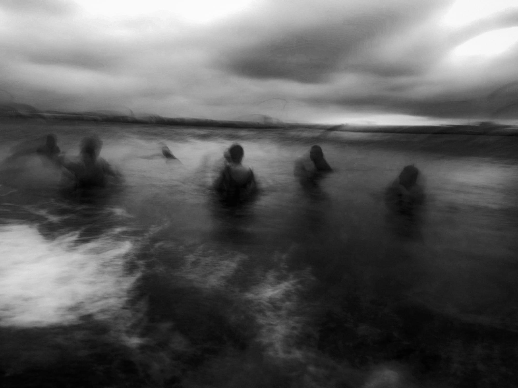 A group of figures wades through a dark, turbulent ocean under a stormy sky. The image is in black and white, creating a moody atmosphere, with blurred details that enhance a sense of motion and mystery. The silhouettes of the individuals are partially submerged, adding to the dramatic feeling of the scene.