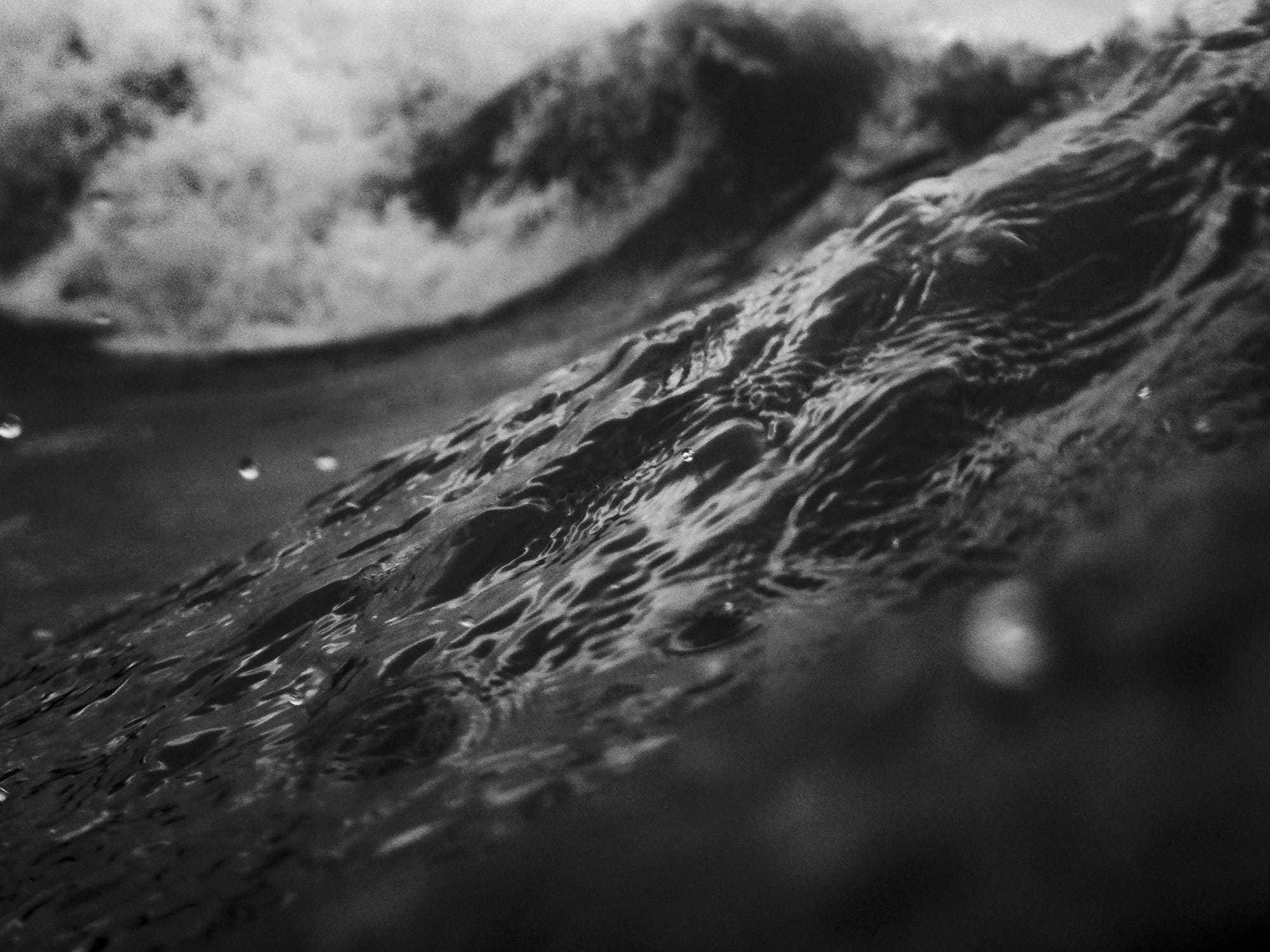 A close-up, monochromatic image of a wave in motion, showcasing the texture and movement of the water. The surface features intricate patterns created by ripples and bubbles, conveying a sense of fluidity and dynamism. The contrasting light and shadow enhance the depth and volume of the wave, evoking a dramatic oceanic atmosphere.