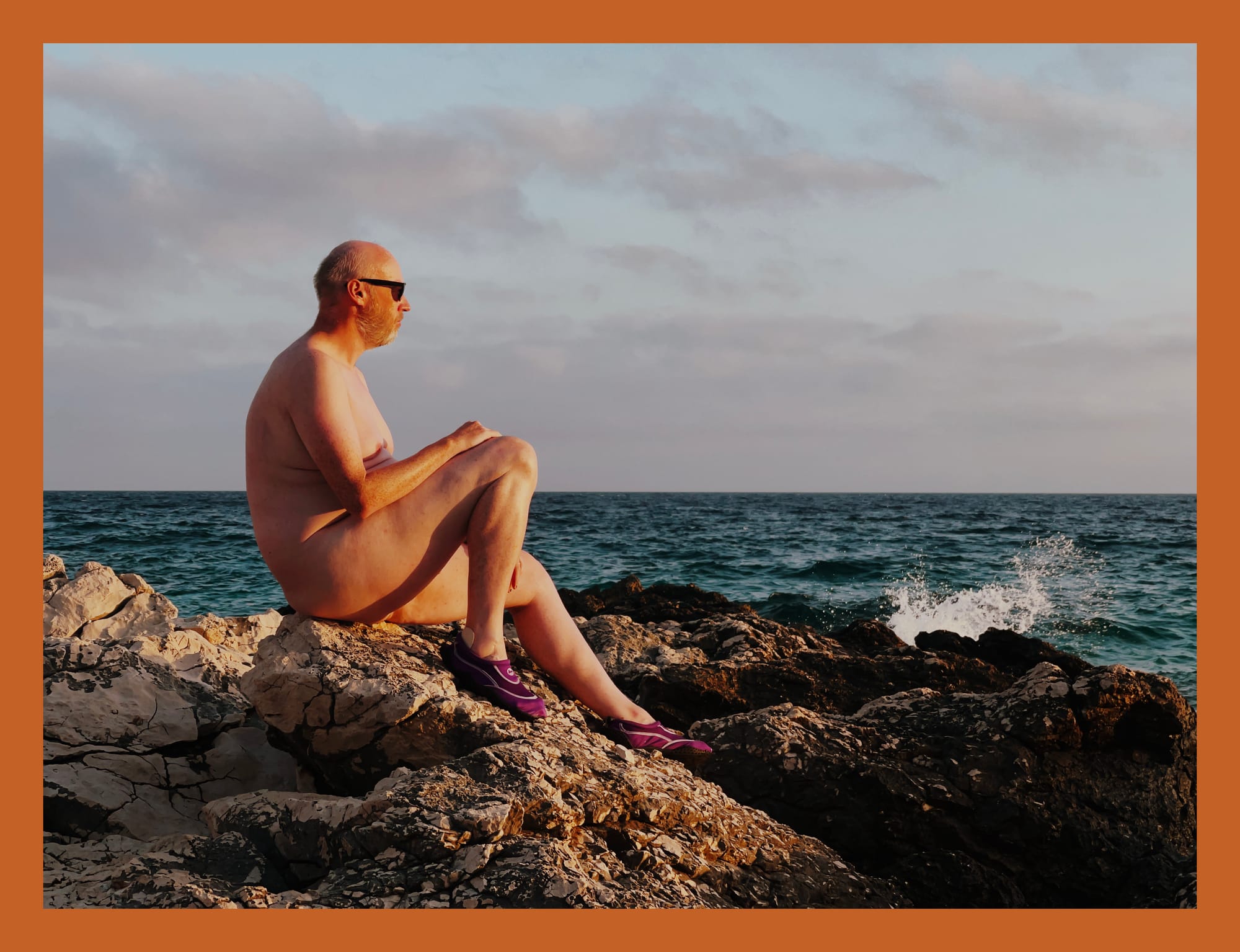 A man sits on a rocky shore, facing the ocean. He is wearing sunglasses and a relaxed expression, while the waves gently crash against the rocks. The sky is partly cloudy, suggesting a serene atmosphere. The image has a warm, orange border framing the scene.