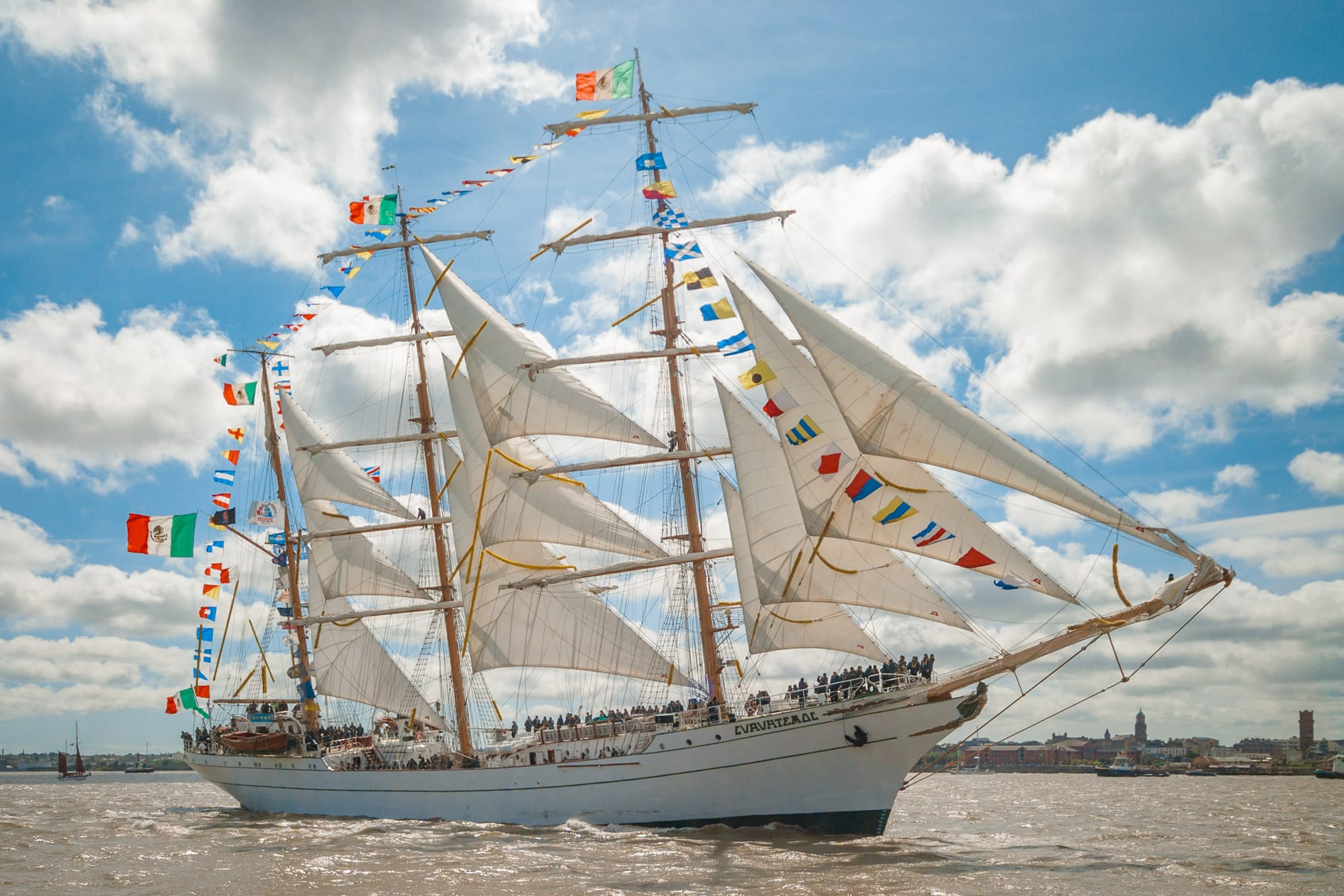 A tall ship, with all the sails unfurled, sails down the River Mersey on a sunny day.