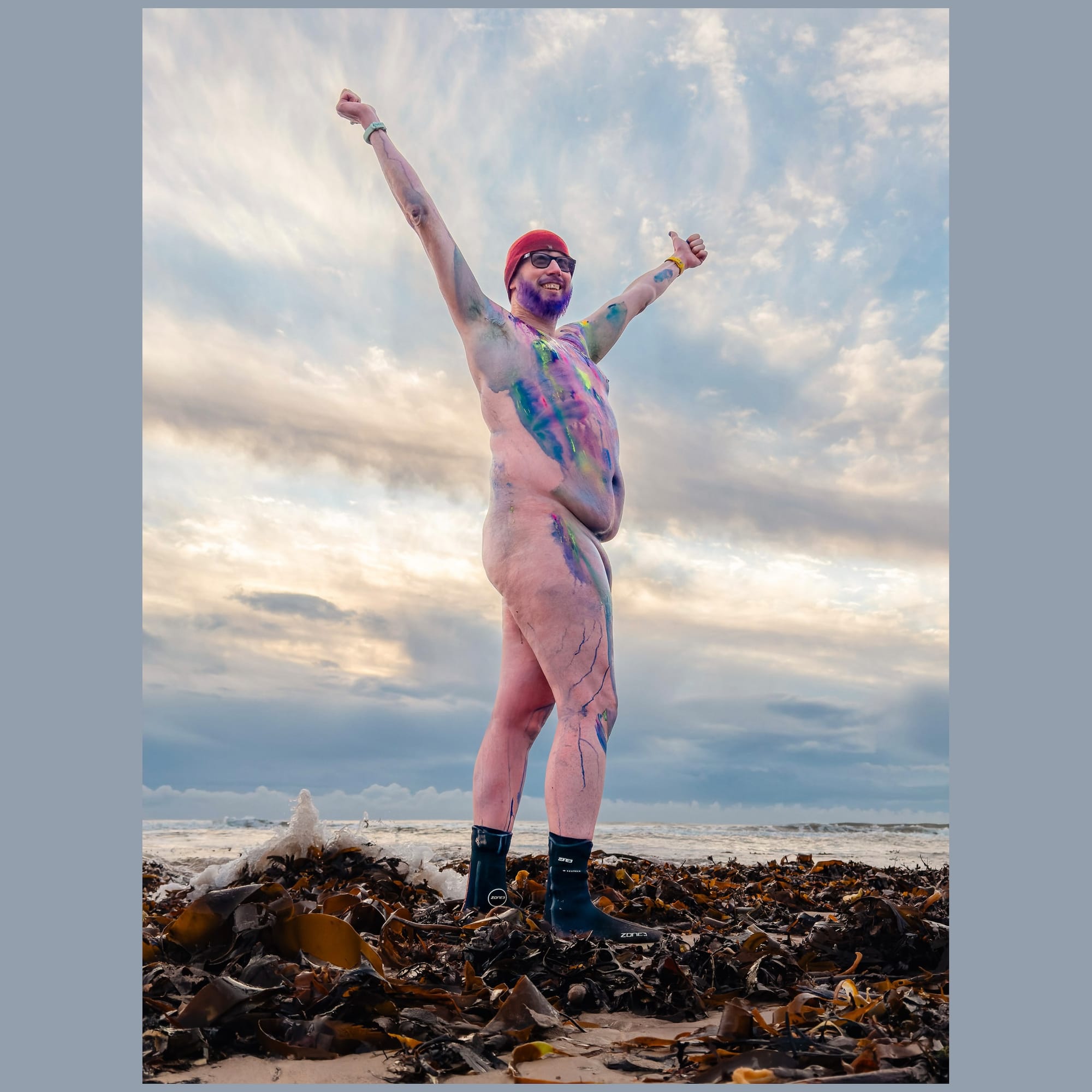 A naked male bodied person smiles and holds up their arms on a beach. They are covered in colourful body paint.