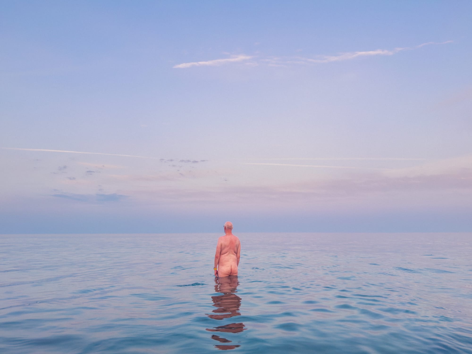 A naked male body stands alone in the sea facing away from the camera. There is a pinkness to the sky at sunrise