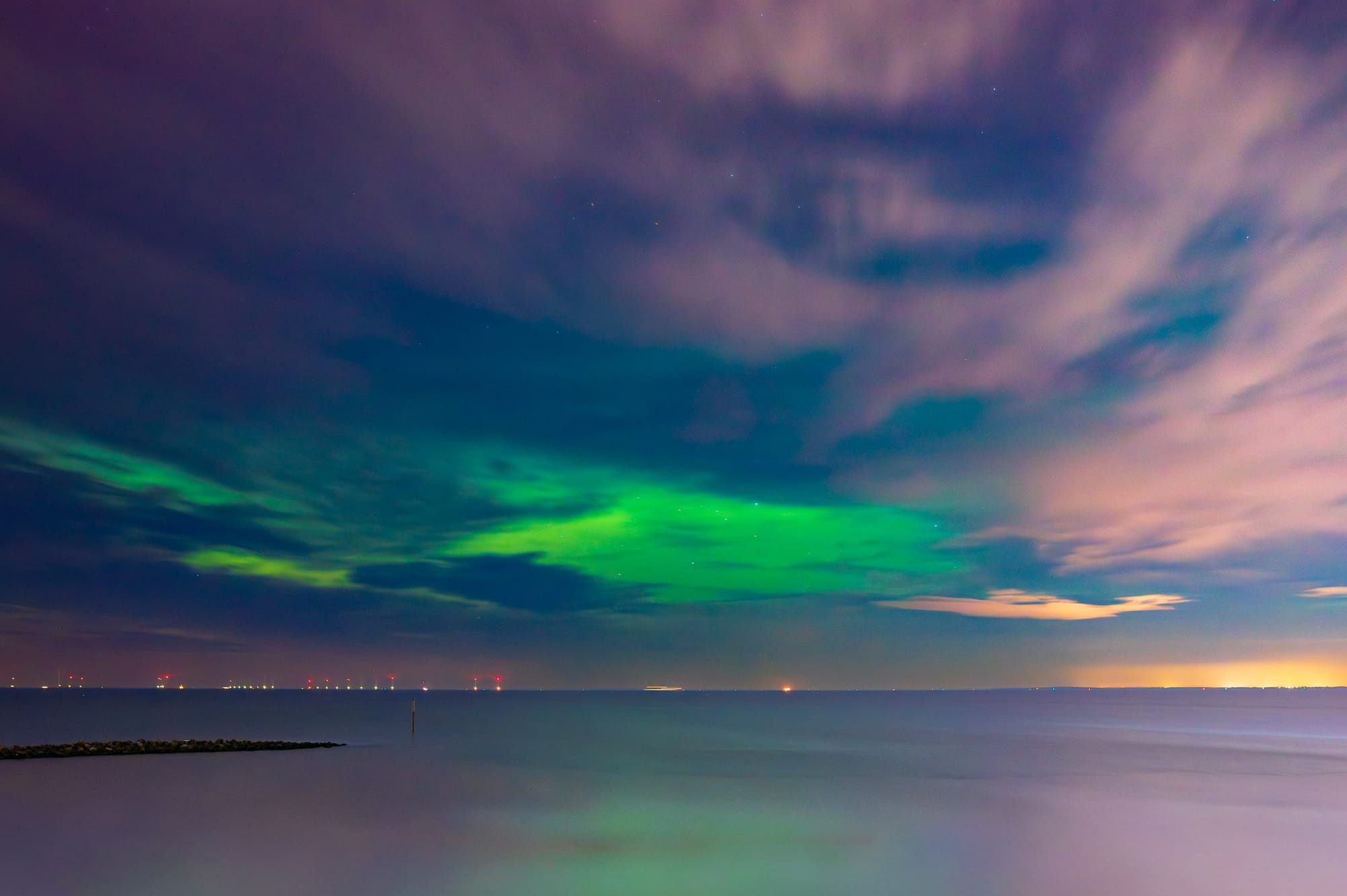 The sky is lit up with a green aurora and pinkish clouds above a calm sea.