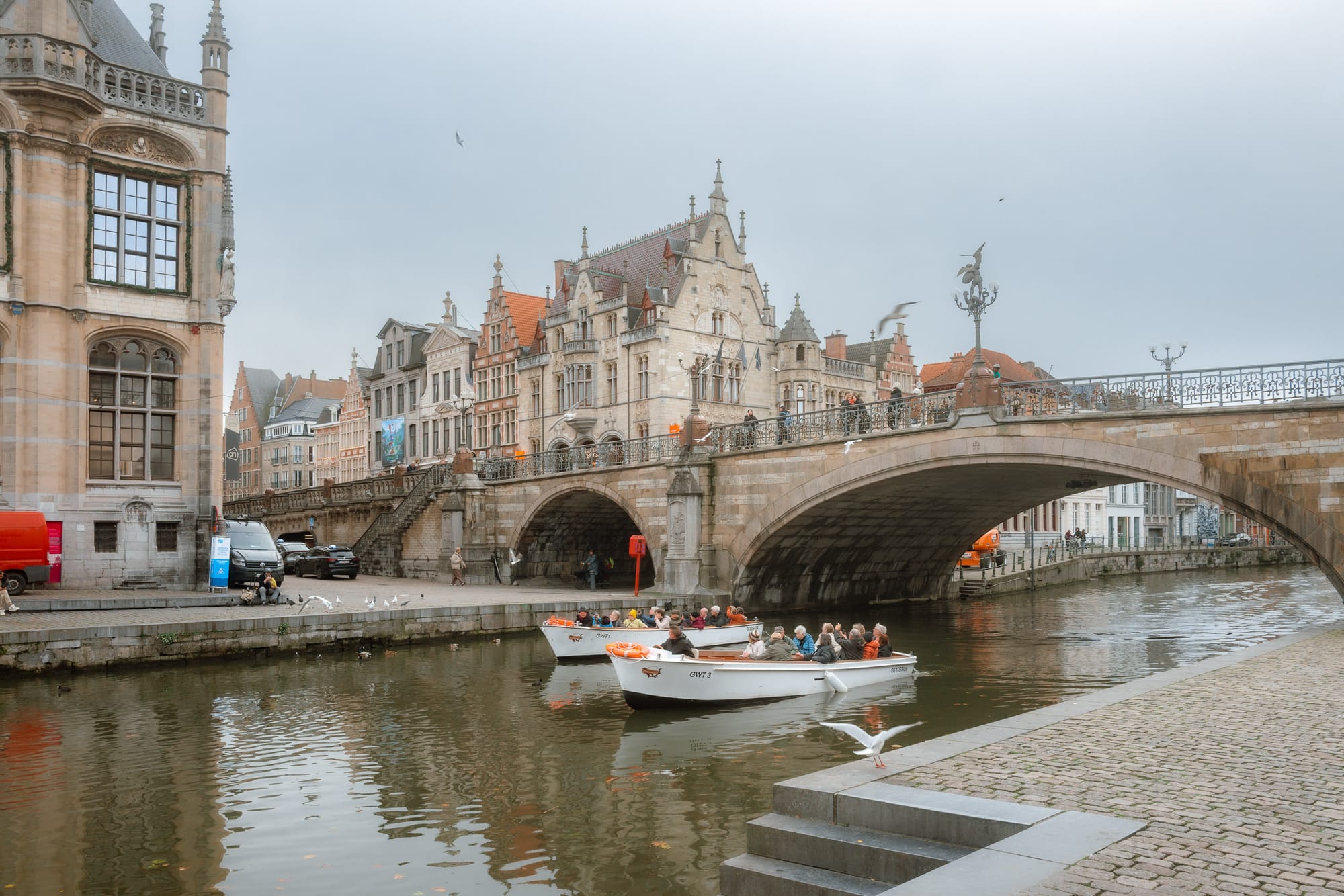 Boats on the canal in Ghent, Belgium, with historic buildings and a bridge in the background.