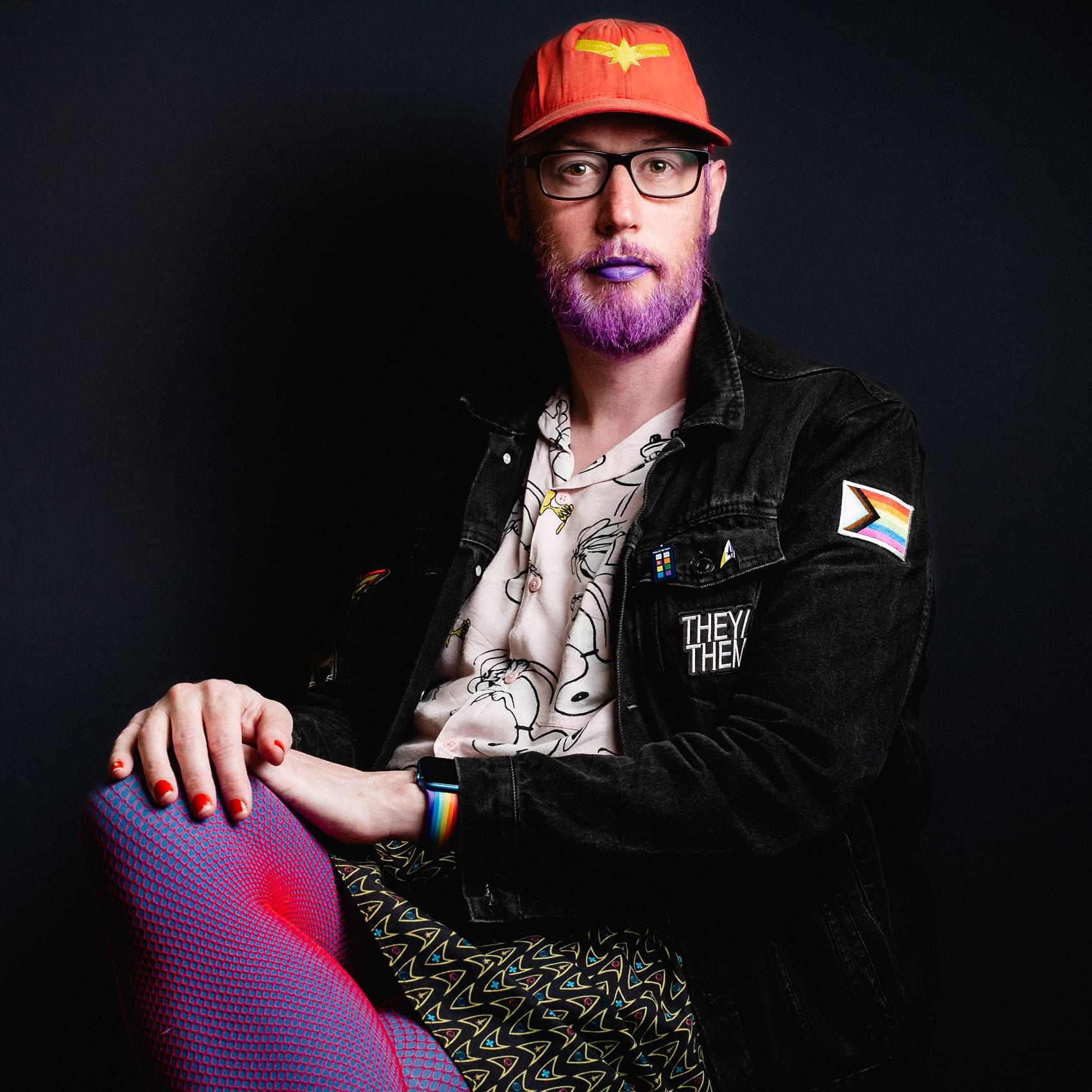 A person seated against a dark background, wearing a red cap and glasses, showcases a vibrant, gender-fluid style with a printed shirt, denim jacket adorned with patches, and colorful patterned leggings. They have brightly colored lipstick and nail polish that complement their unique outfit.
