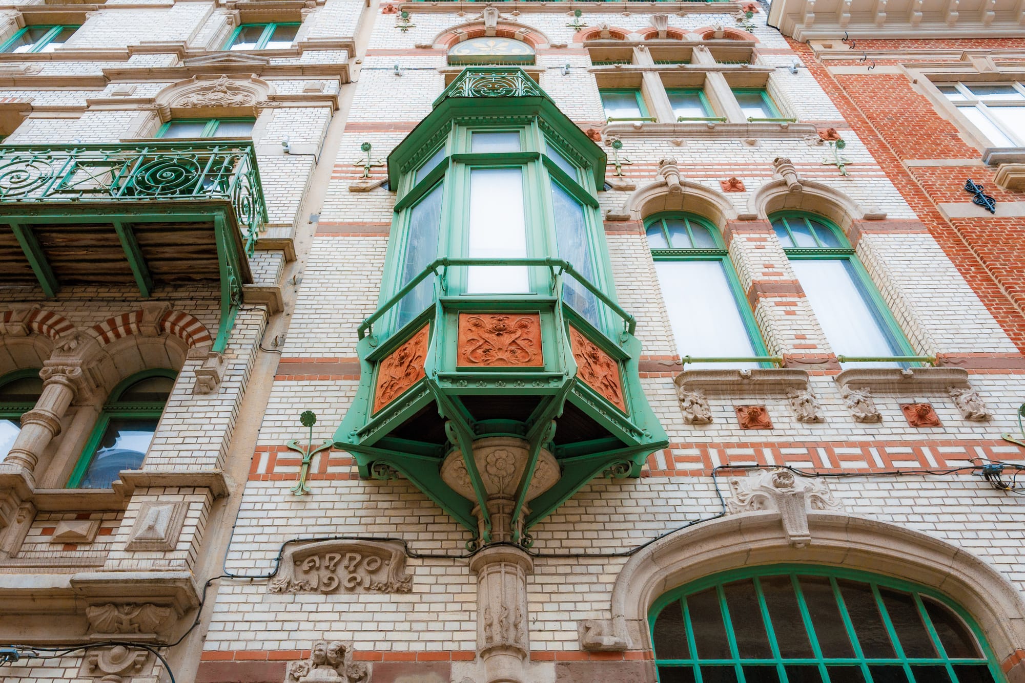 A close-up view of an ornate architectural feature on a historic building, showcasing a green and orange bay window with intricate decorative elements. The surrounding facade is a mix of white brick and red accents, highlighting elegant detail and craftsmanship. Green balconies and window frames complement the color scheme, contributing to the building's unique character.
