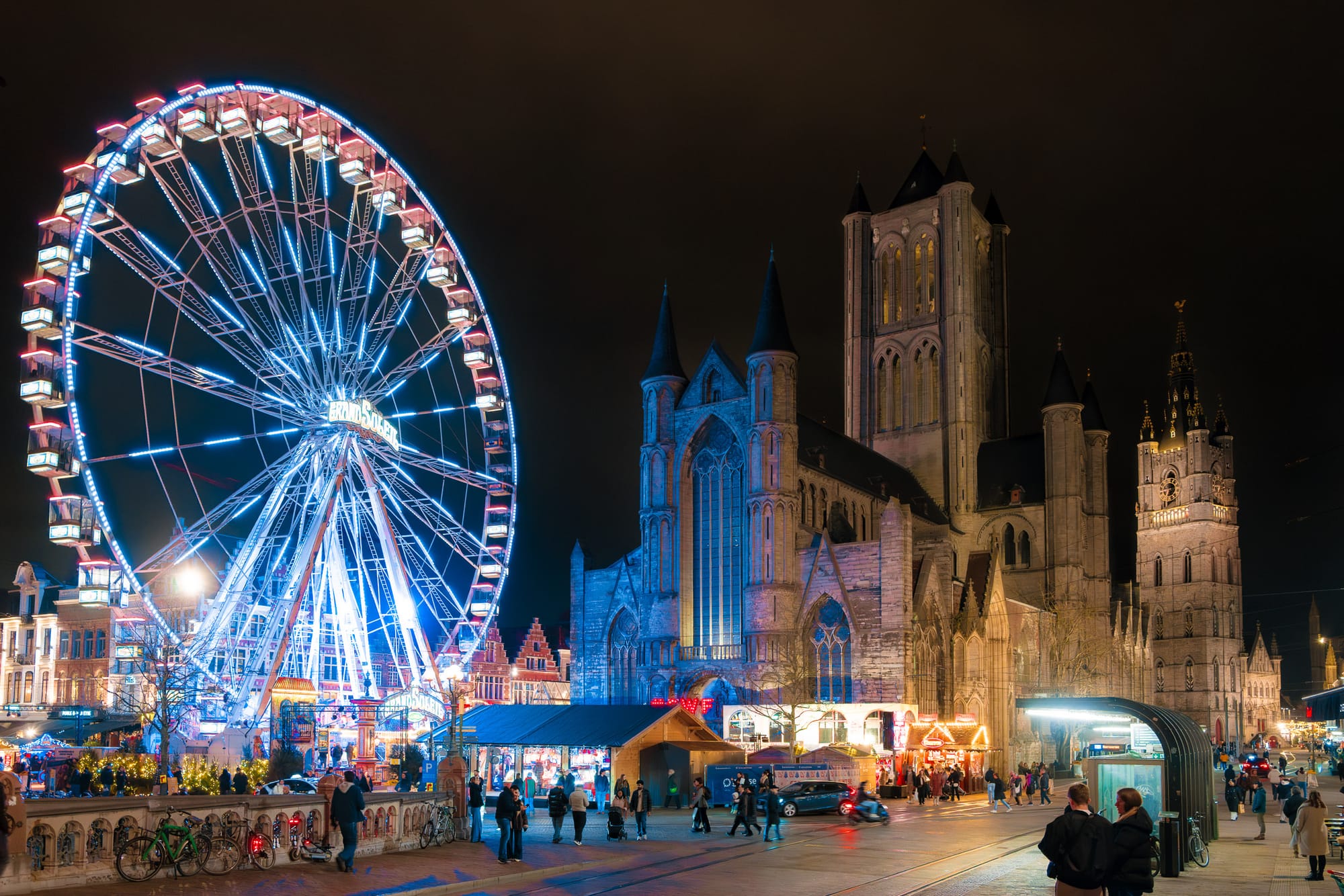 A vibrant night scene showcasing a large illuminated Ferris wheel beside a historic Gothic cathedral. The Ferris wheel is adorned with colorful lights, creating a festive atmosphere. People can be seen enjoying the fairground activities, and the surrounding area features charming buildings and stalls, enhancing the lively ambiance of the setting.