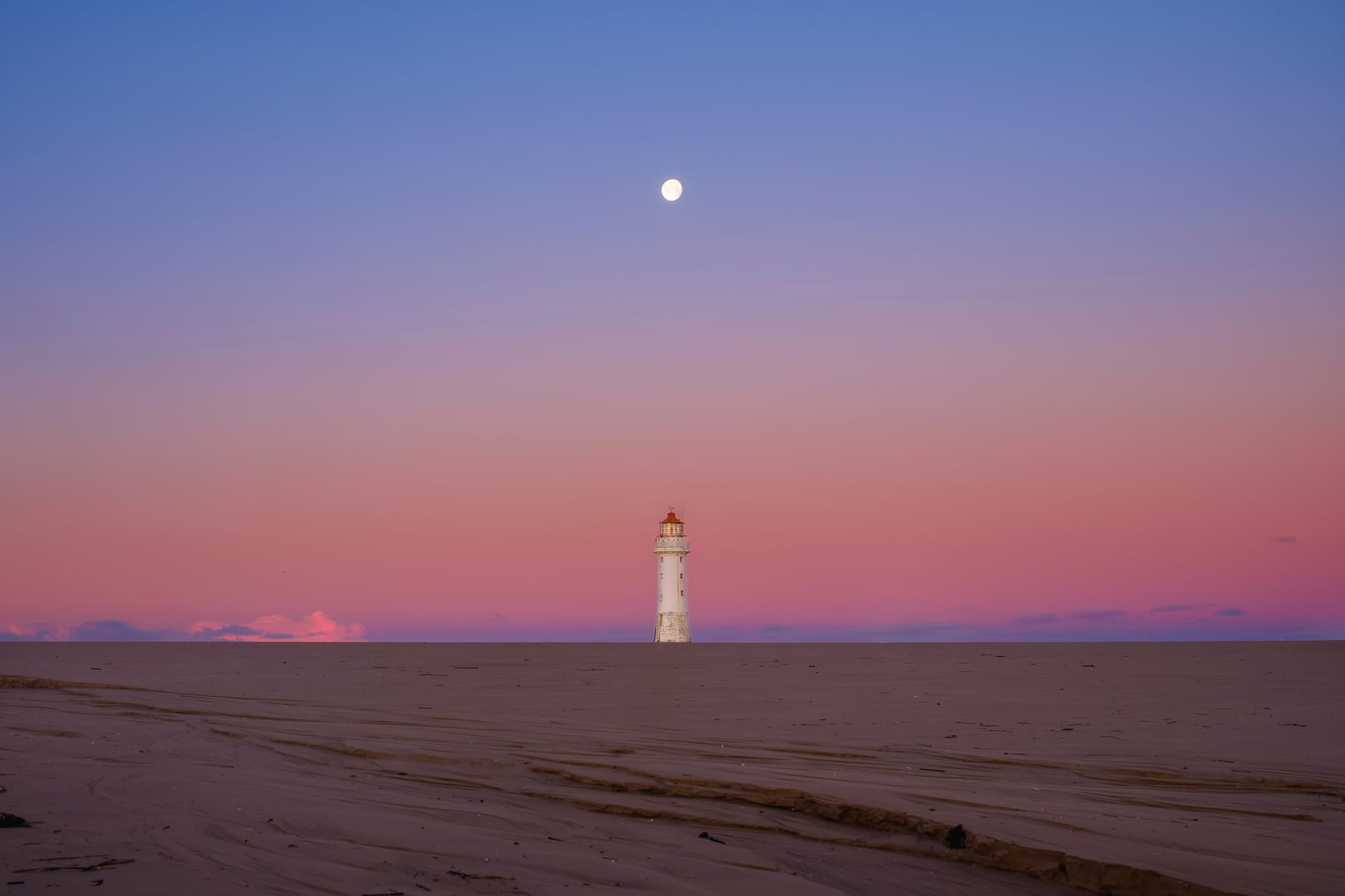 The moon sets directly over the top of a white lighthouse set against a pink sky on a beach.