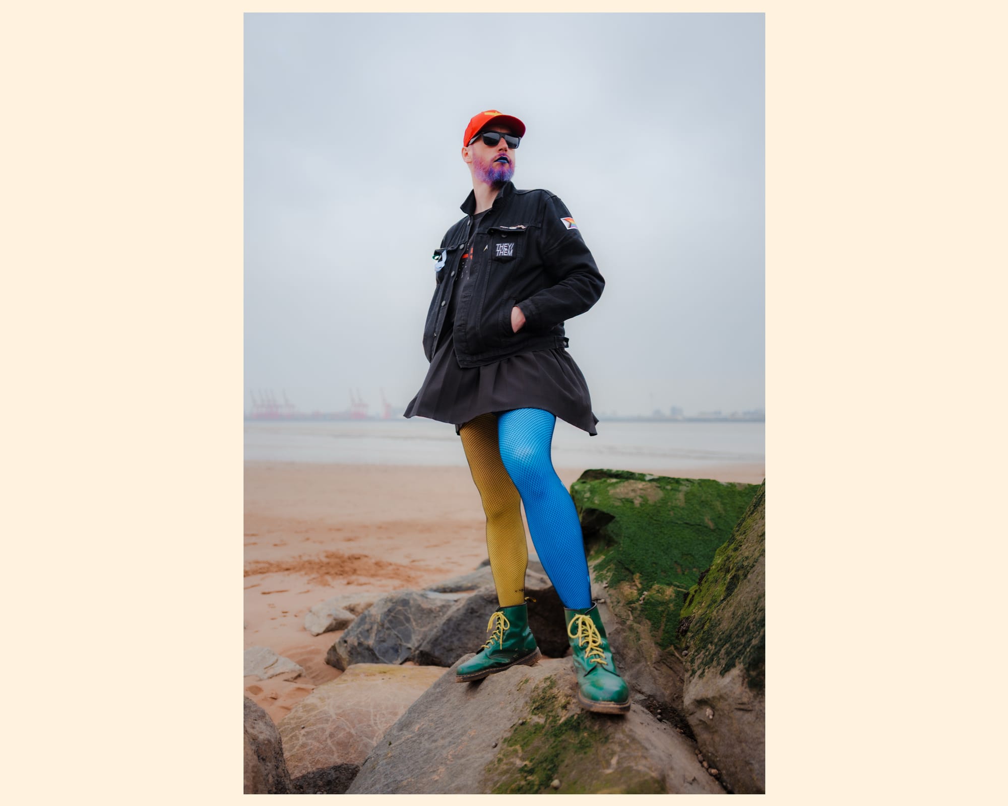 A person stands confidently on rocky terrain near a body of water, dressed in a black jacket and a short skirt. They wear striking blue and yellow leggings, coupled with green boots. Their outfit is completed with an orange cap, and they have vivid purple facial hair, creating a bold and unique fashion statement against a cloudy sky backdrop.