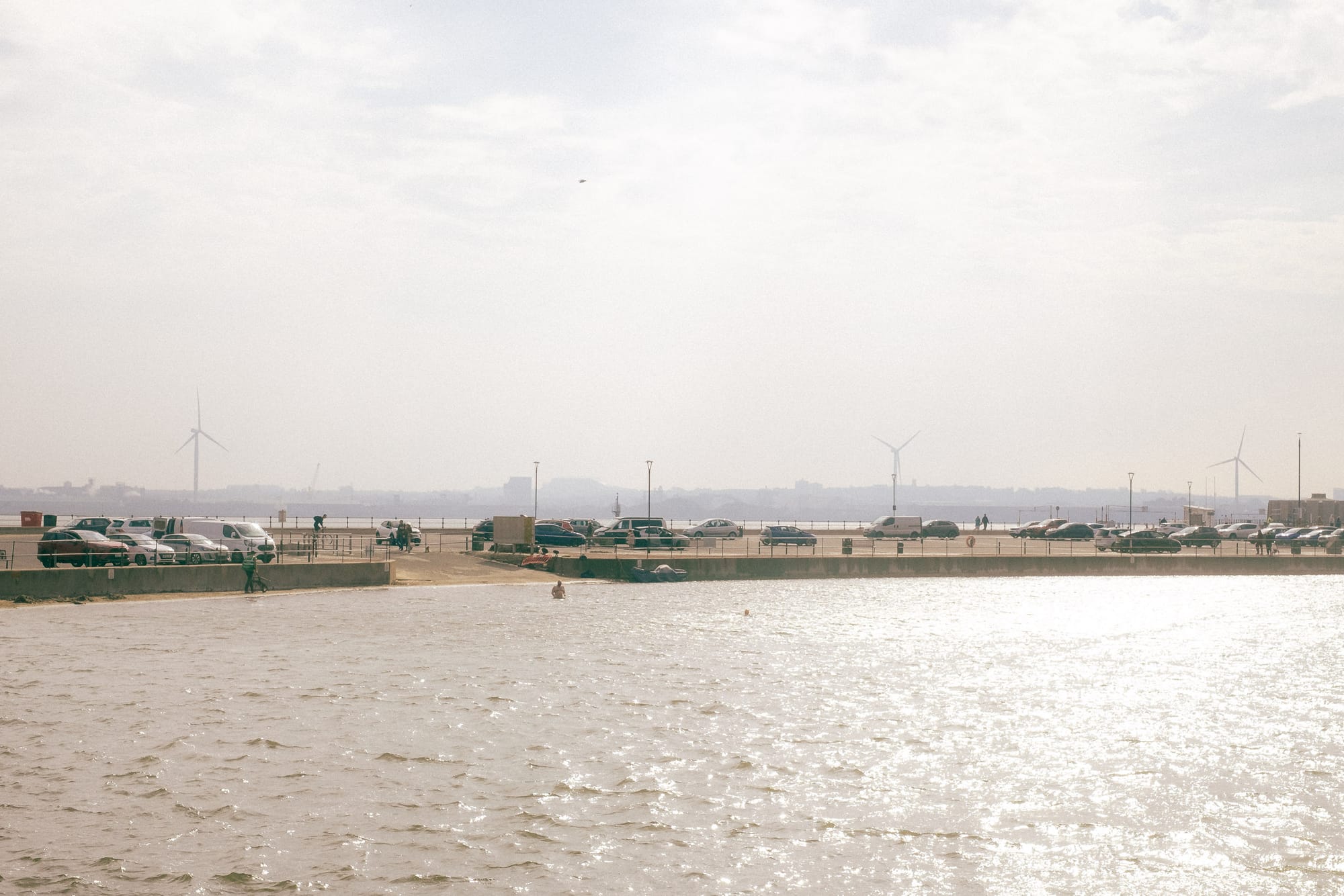 A wide shot of a coastal scene with a concrete pier, parked cars, and wind turbines in the background under a cloudy sky.